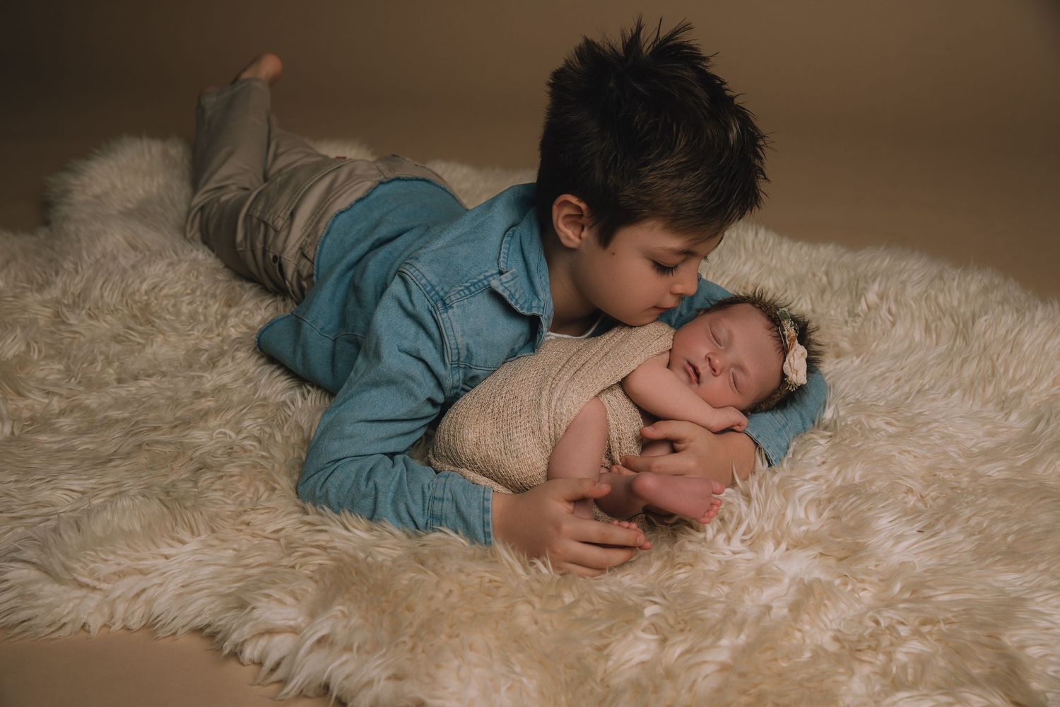Older boy in blue shirt kisses sleeping newborn baby sister wrapped in white on a fluffy cream rug.