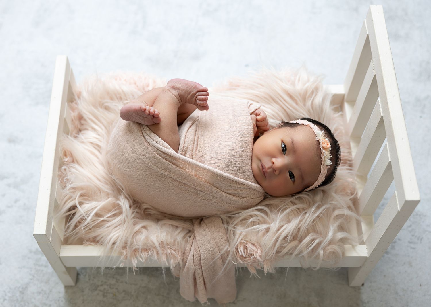 Newborn baby girl in pink wrap resting on fluffy fur in a white miniature bed for a professional photo.