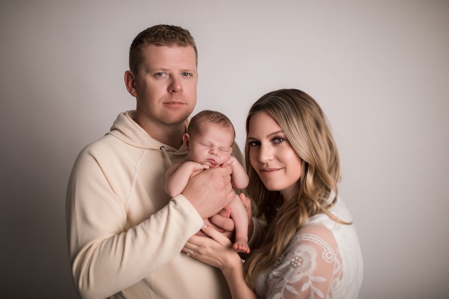 A couple in a photography studio holding their sleeping newborn baby, dressed in cream and white outfits.