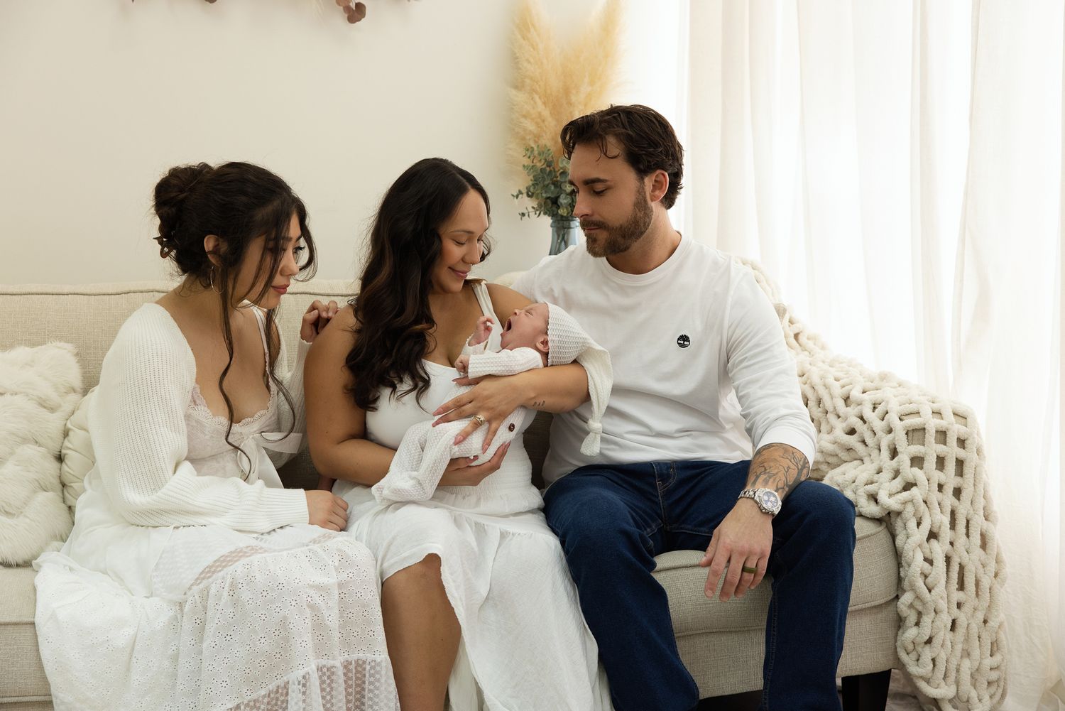 Family gathered on a couch admiring a newborn baby, with two women in white dresses and a man in a white shirt.