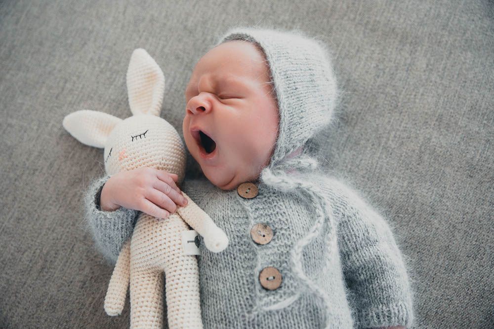 Sleeping newborn baby in gray knit outfit yawning while holding a crocheted bunny stuffed animal in the Bonsall Studio near Fallbrook, Vista