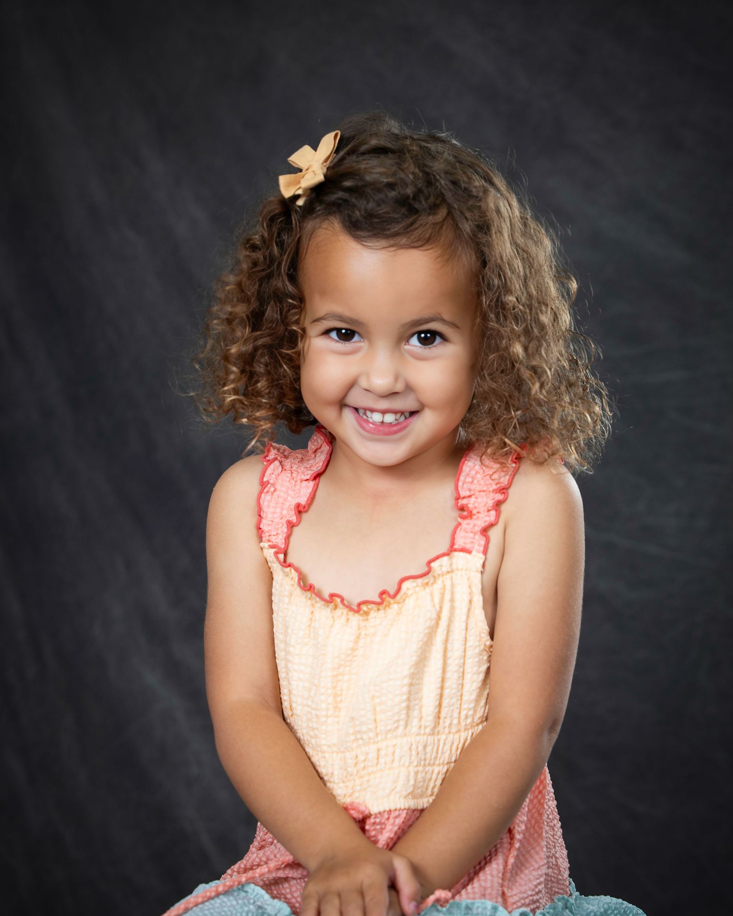 A child wearing a peach-colored dress with ruffles poses against a dark backdrop in a studio portrait school session in the Bonsall Studio n