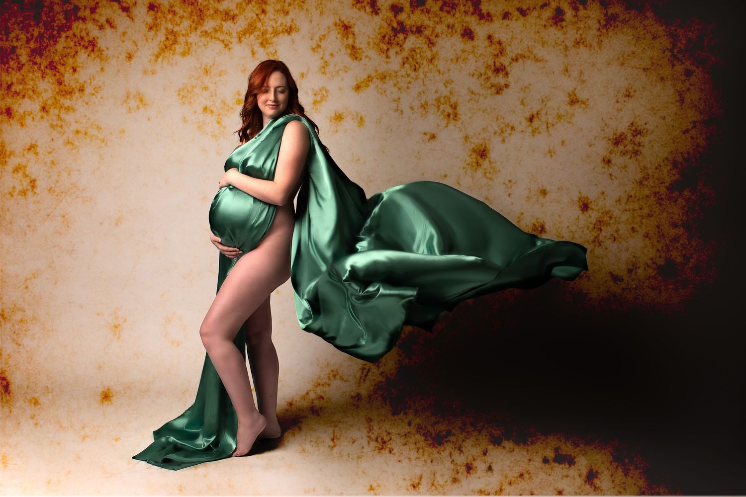 Pregnant woman draped in flowing green satin fabric against a rustic backdrop for a maternity photo shoot. Bonsall, near Fallbrook, & Temecu