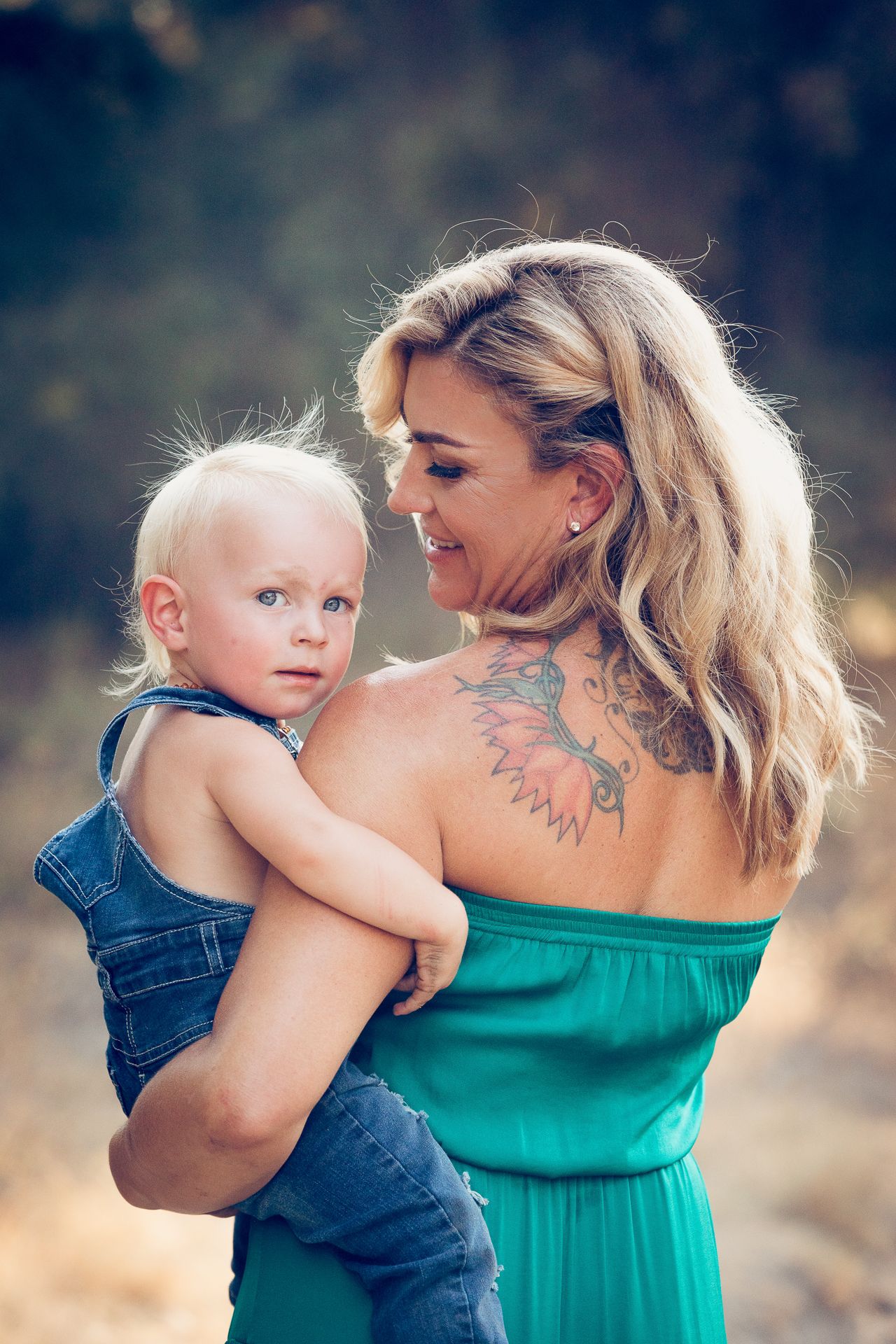 Blonde mother in green dress holding toddler outdoors, showcasing shoulder tattoo in Fallbrook CA