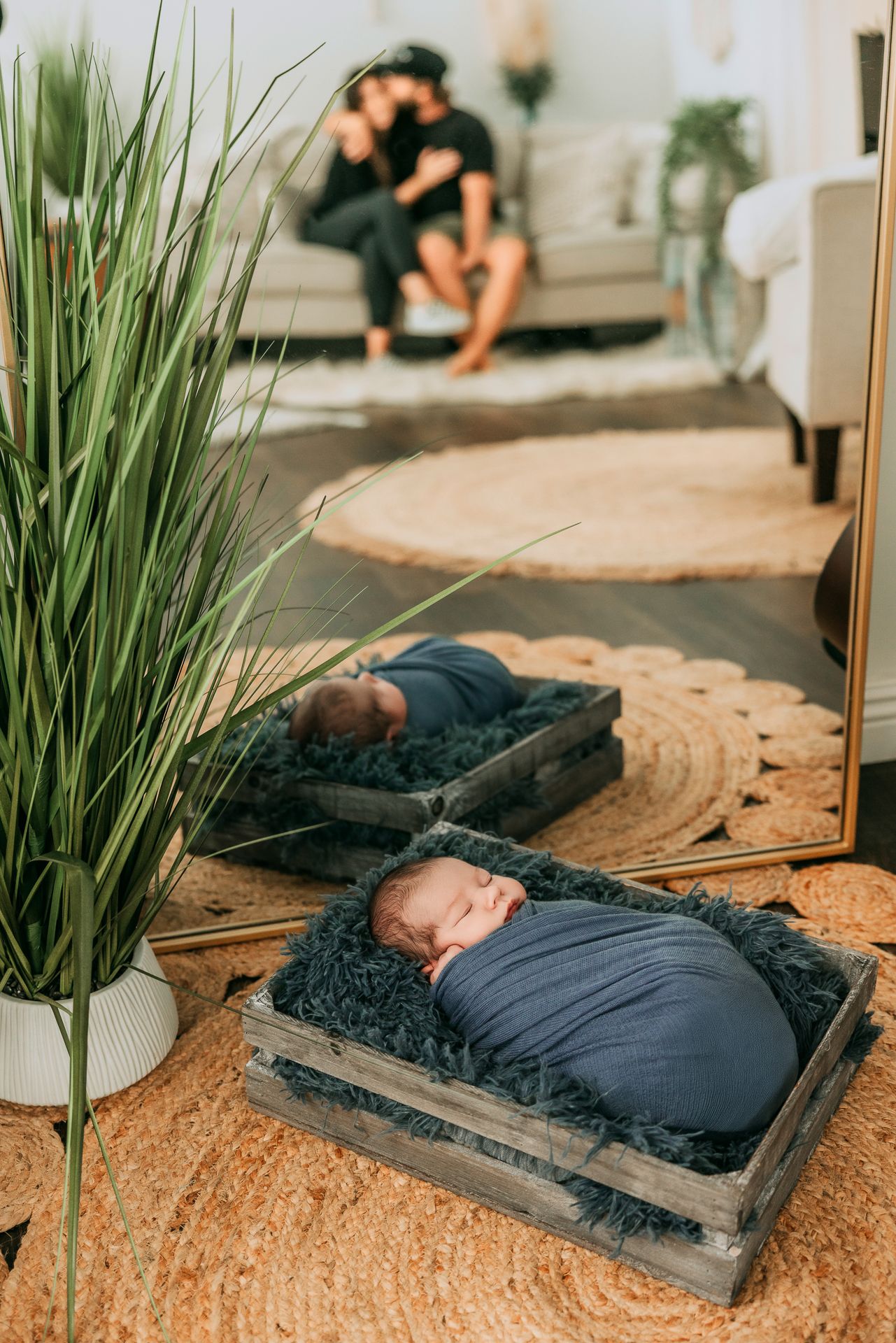 Sleeping newborn baby swaddled in navy wrap in wooden crate with mirror reflection of parents.