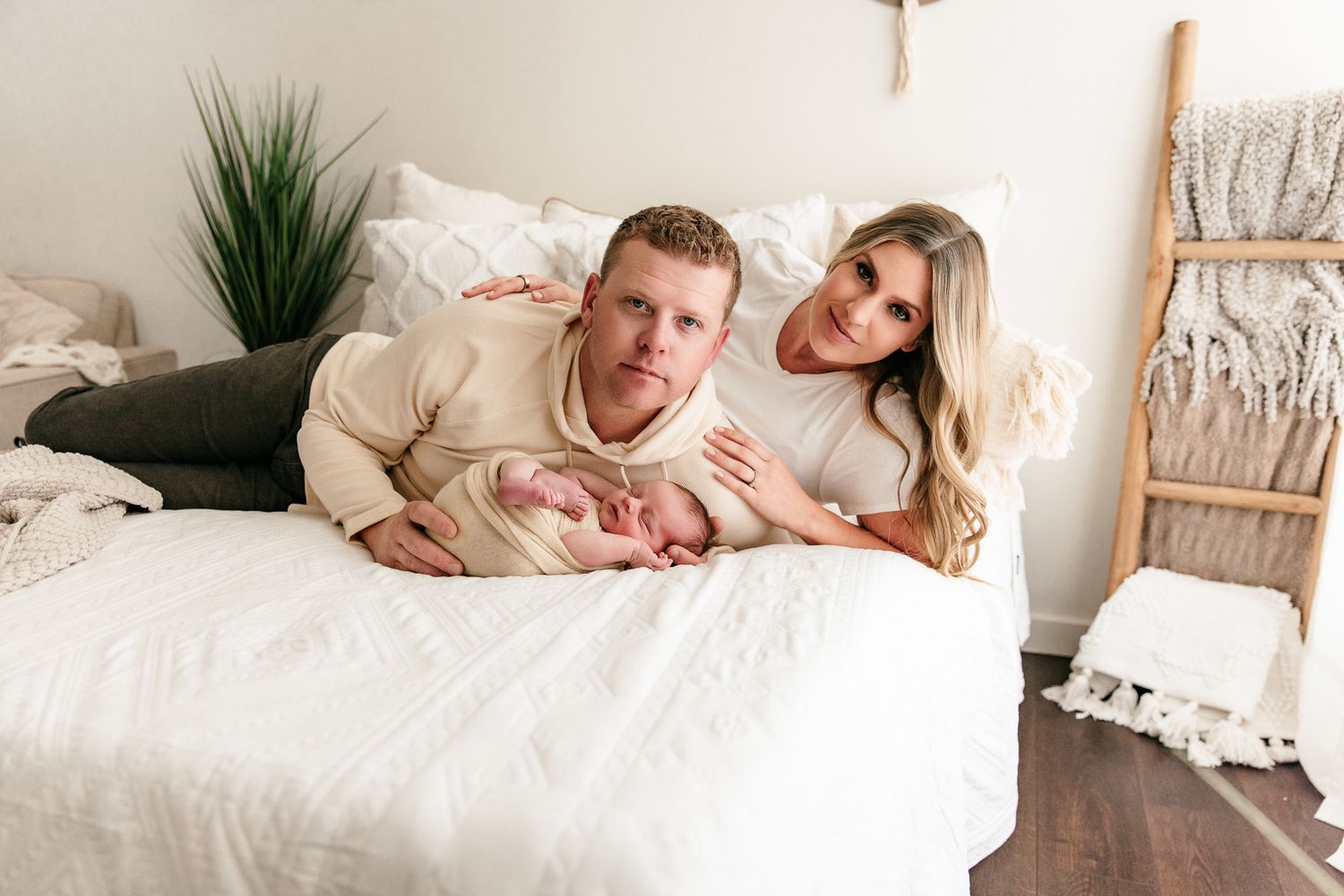 A couple lies on a white bed smiling, cradling their newborn baby in a bright, cozy room with neutral decor.
