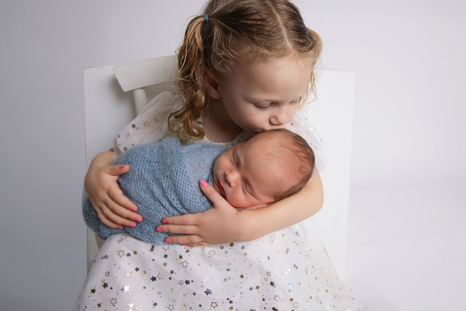 Young girl in star dress gently holds and kisses sleeping newborn baby wrapped in blue swaddle.