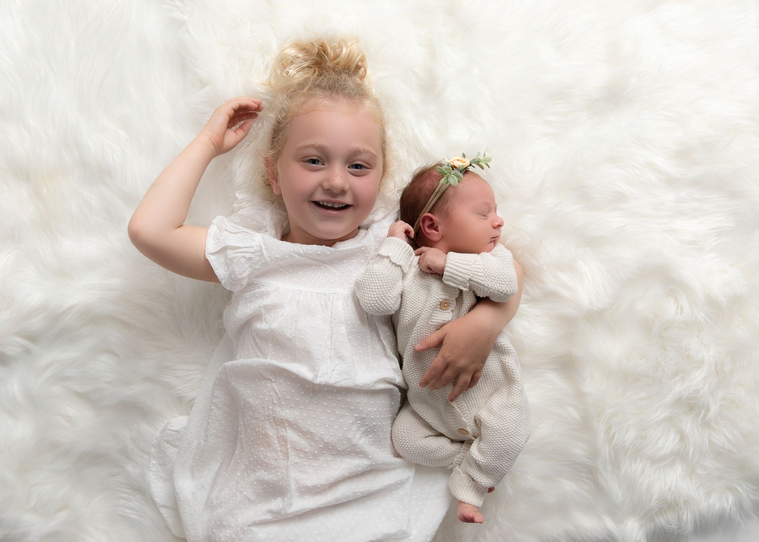 Young girl with blonde hair smiles beside sleeping newborn baby on white fluffy blanket.
