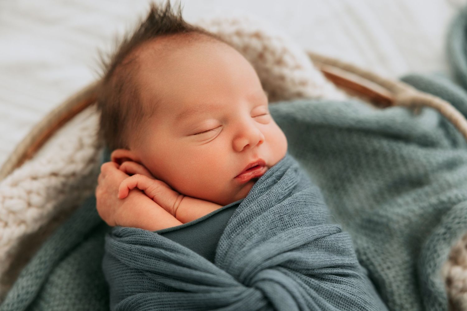 Newborn baby swaddled in teal wrap sleeping peacefully in woven basket during professional newborn photo session.