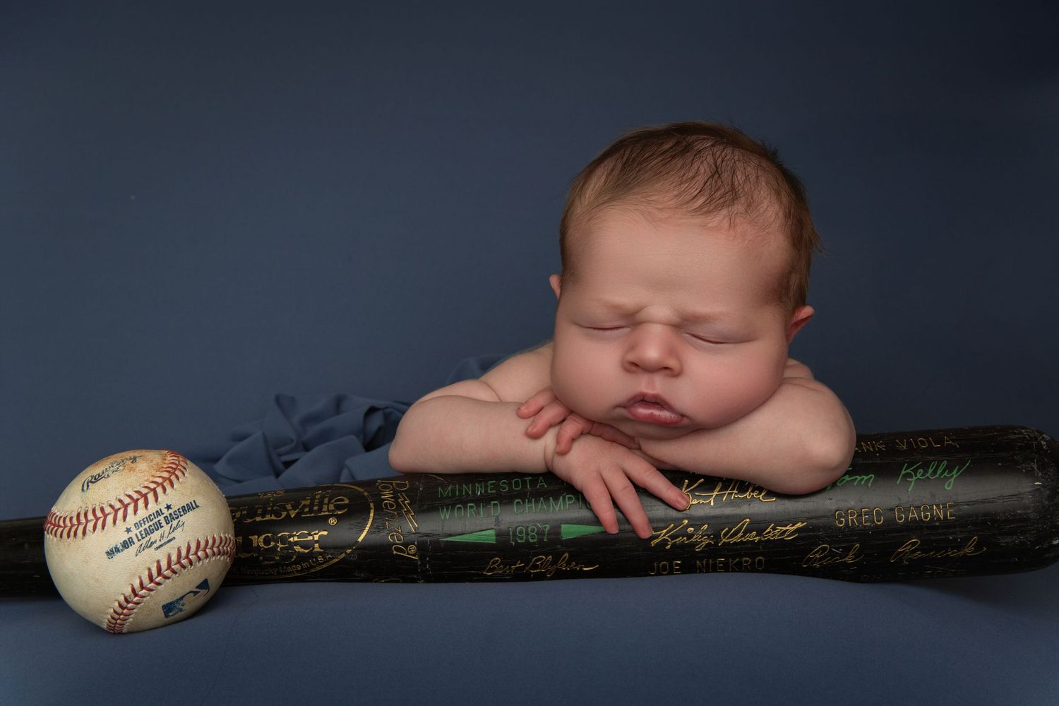 Newborn baby sleeping peacefully on a baseball bat beside a signed baseball against a blue background.