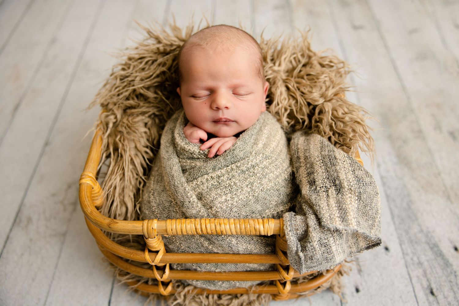 Newborn baby swaddled in a beige knit wrap sleeping peacefully in a wicker basket with fluffy fur lining.