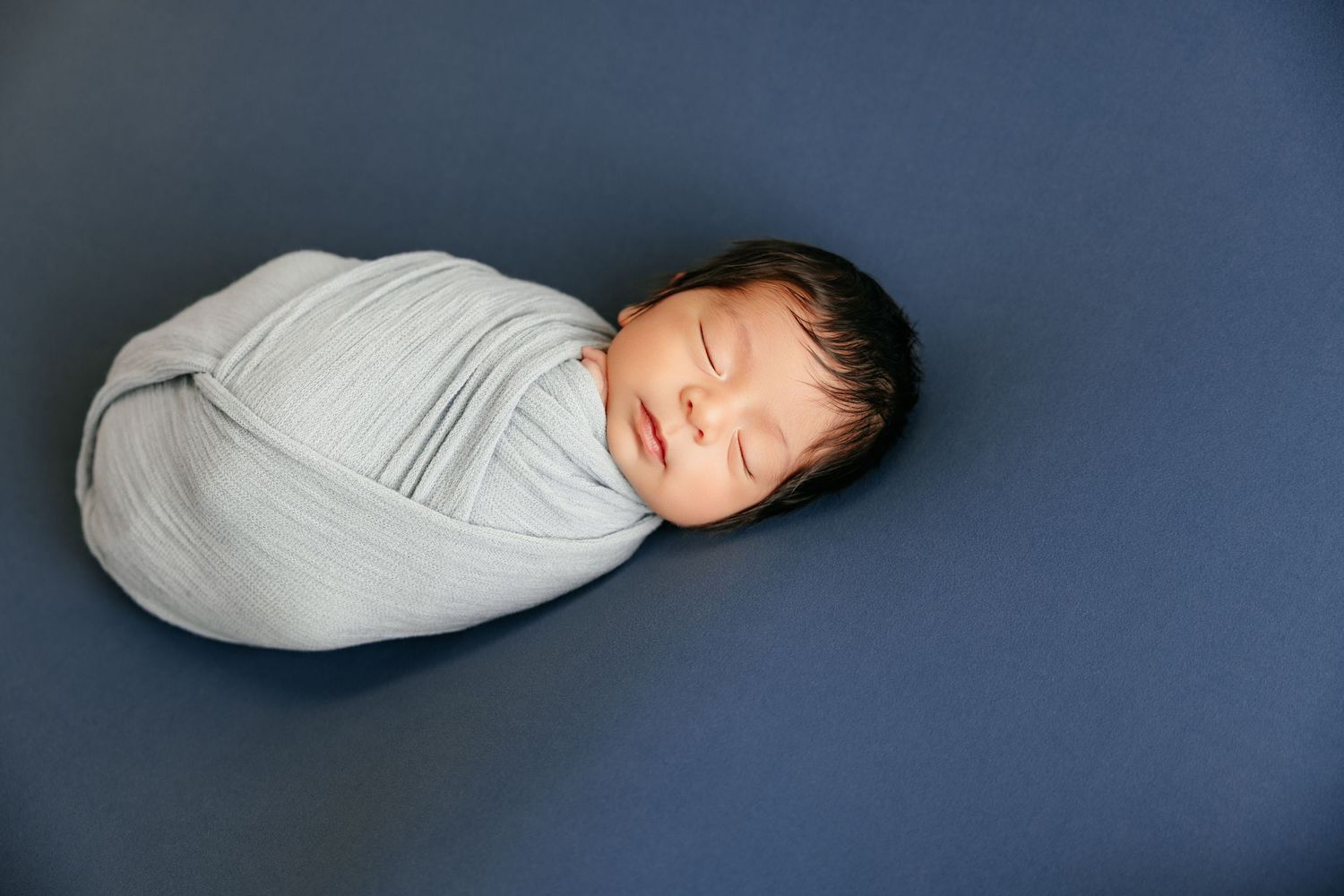 Newborn baby sleeping peacefully swaddled in a gray wrap on a blue background.