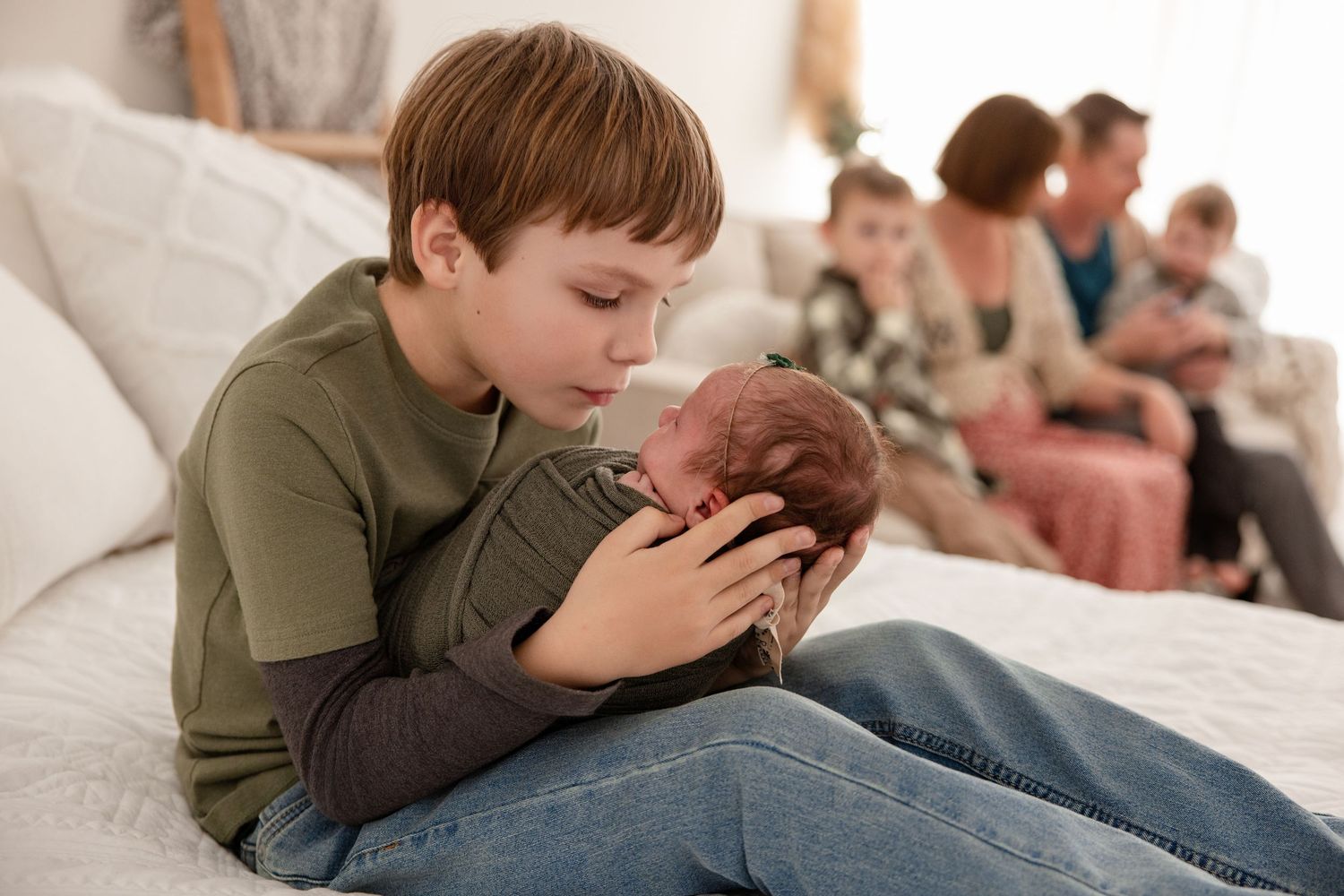A young boy gently holds a sleeping newborn baby while family members sit together in a bright bedroom.