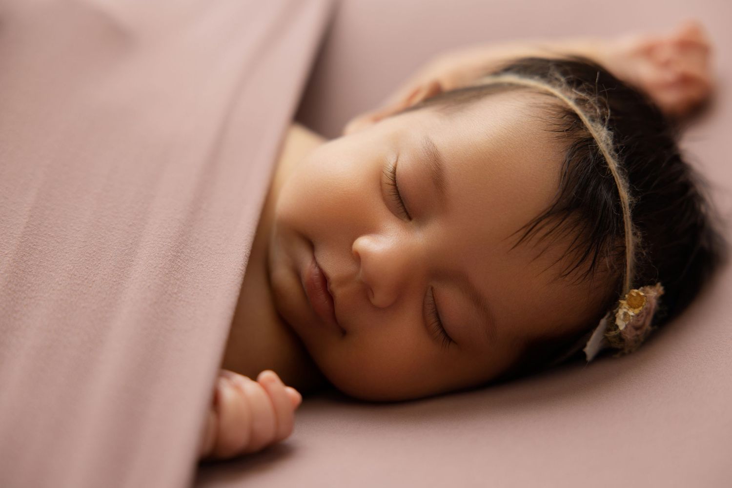 Peaceful sleeping newborn baby girl with dark hair and floral headband on a pink backdrop.