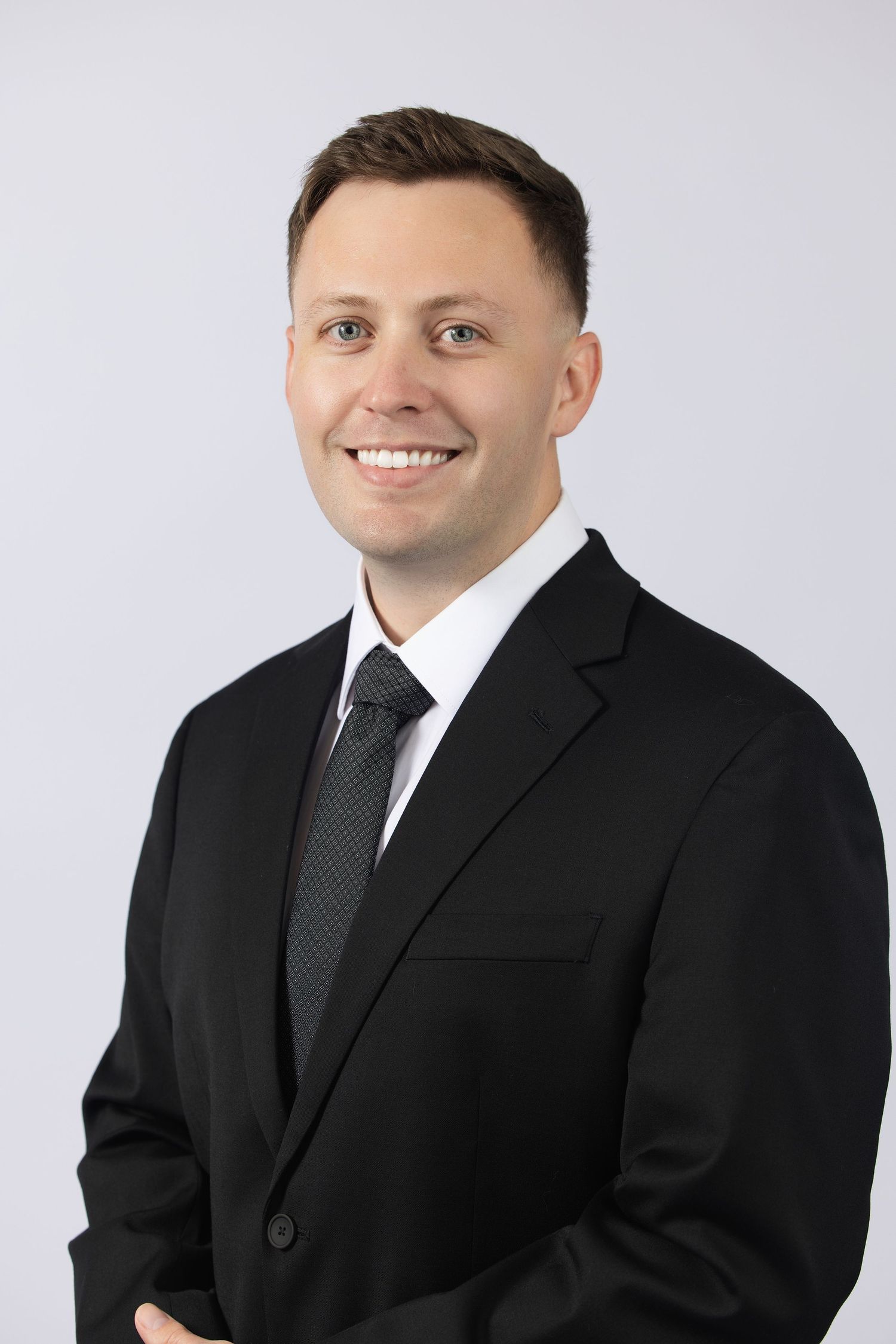 Young professional man in black suit and tie smiling confidently against a light gray background.