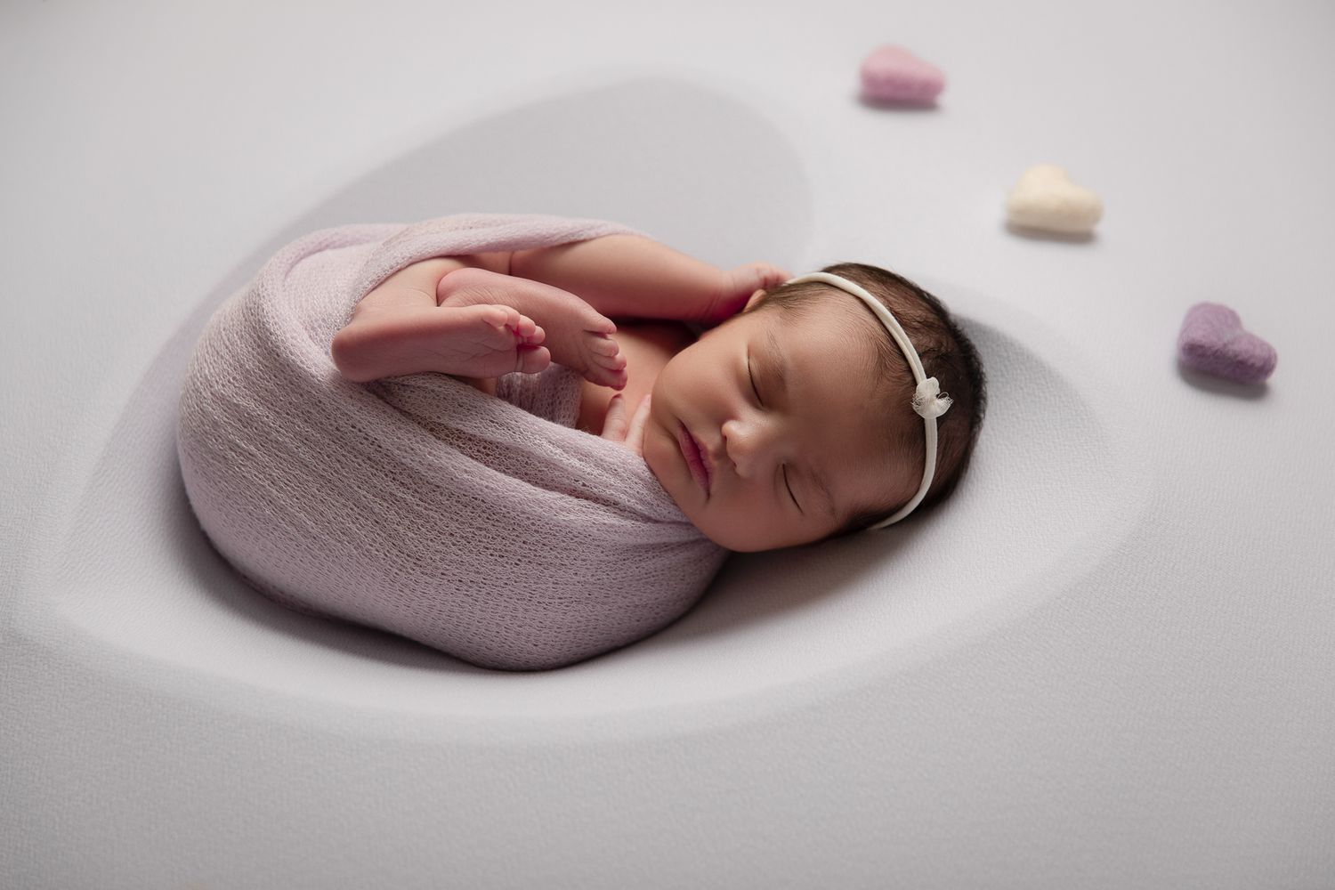 Sleeping newborn baby swaddled in pink wrap posed in heart-shaped bowl with candy hearts nearby at The Studio Bonsall, near Fallbrook/Vista