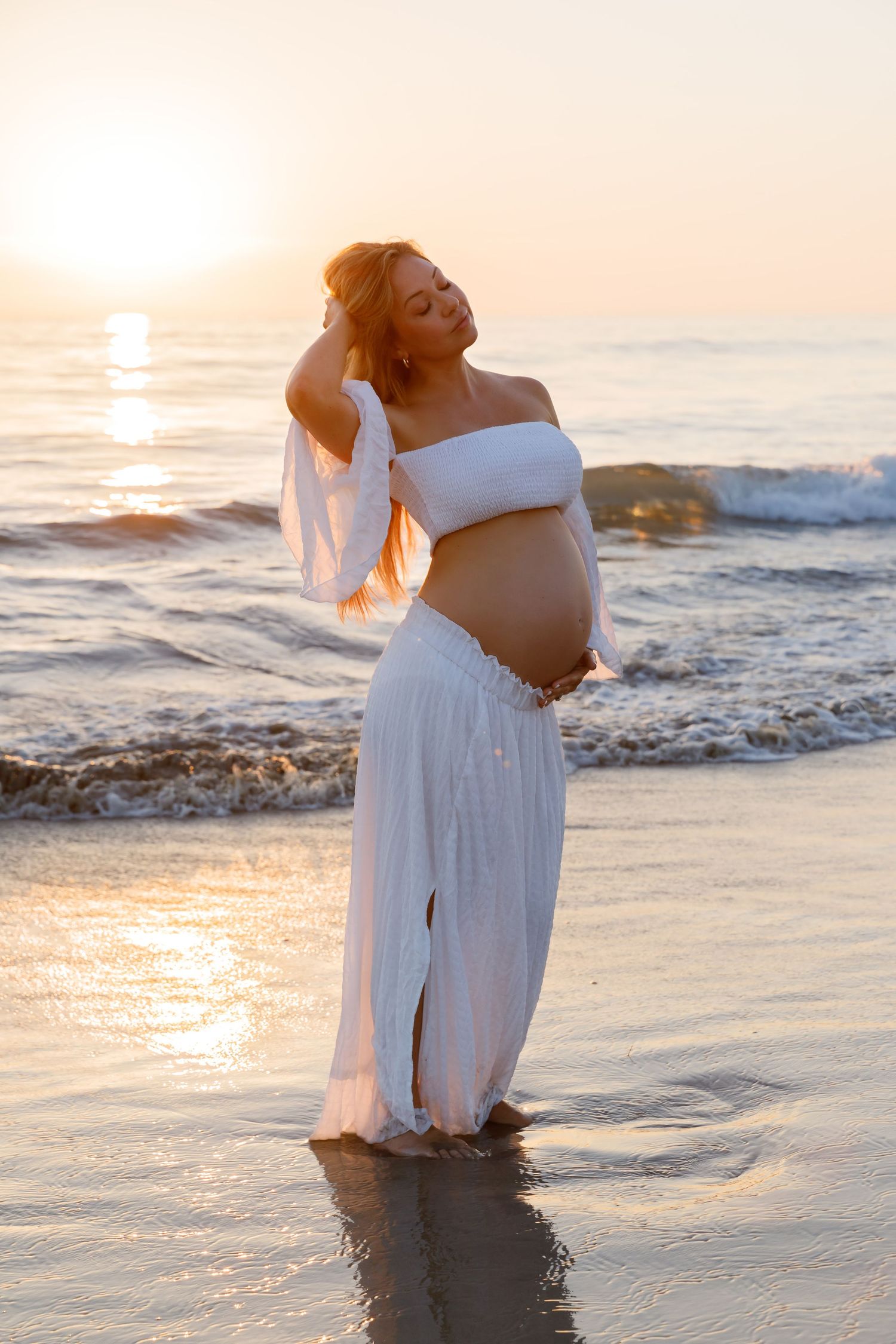 Pregnant woman in white crop top and skirt standing in ocean waves at golden sunset.