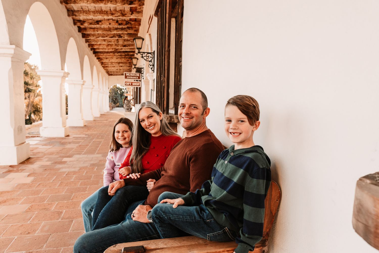 A family of four sits on a bench along a white archway corridor, smiling warmly together.