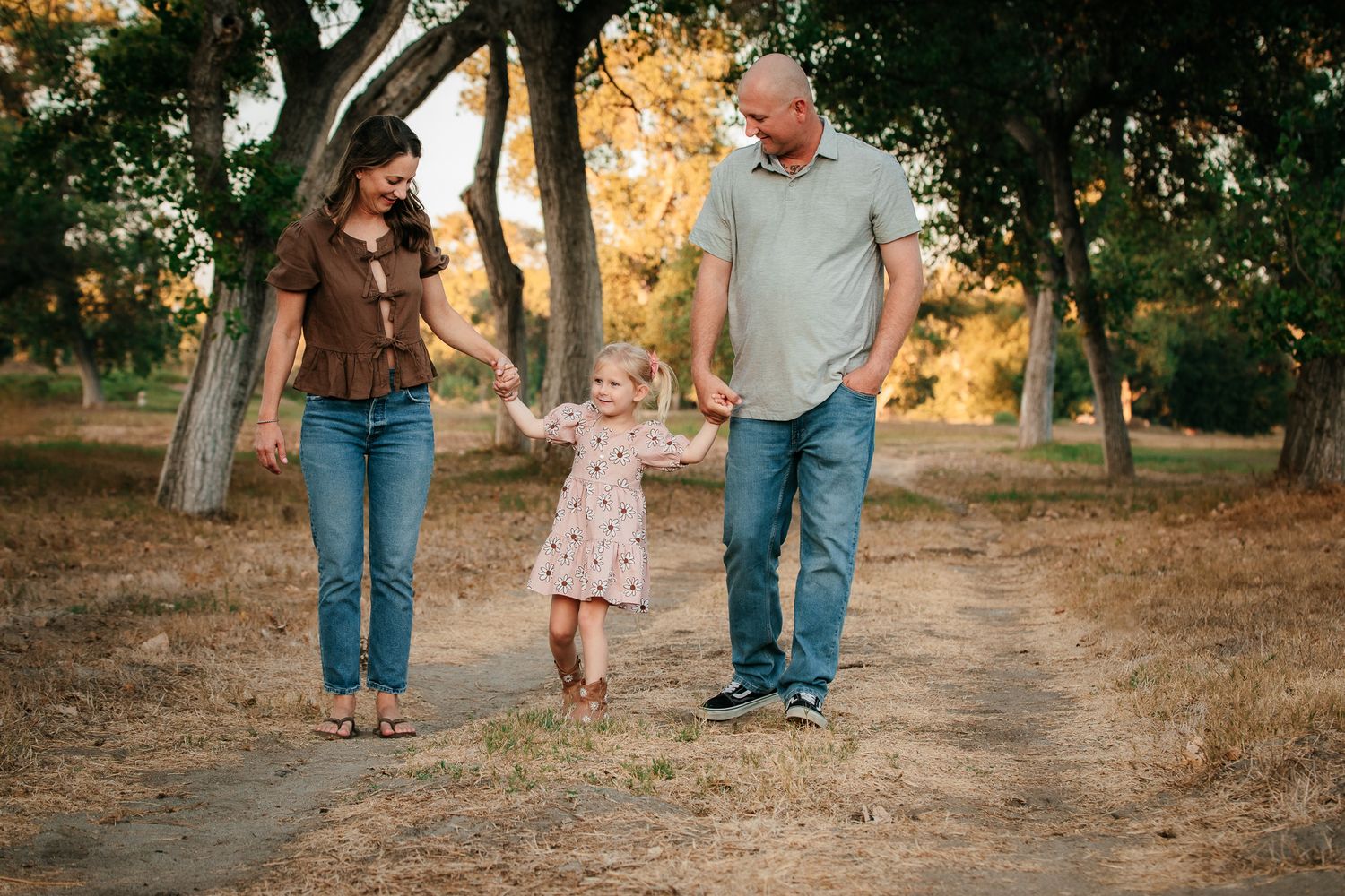 Happy couple swings toddler girl between them on a dirt path surrounded by autumn trees. In Bonsall, near Oceanside