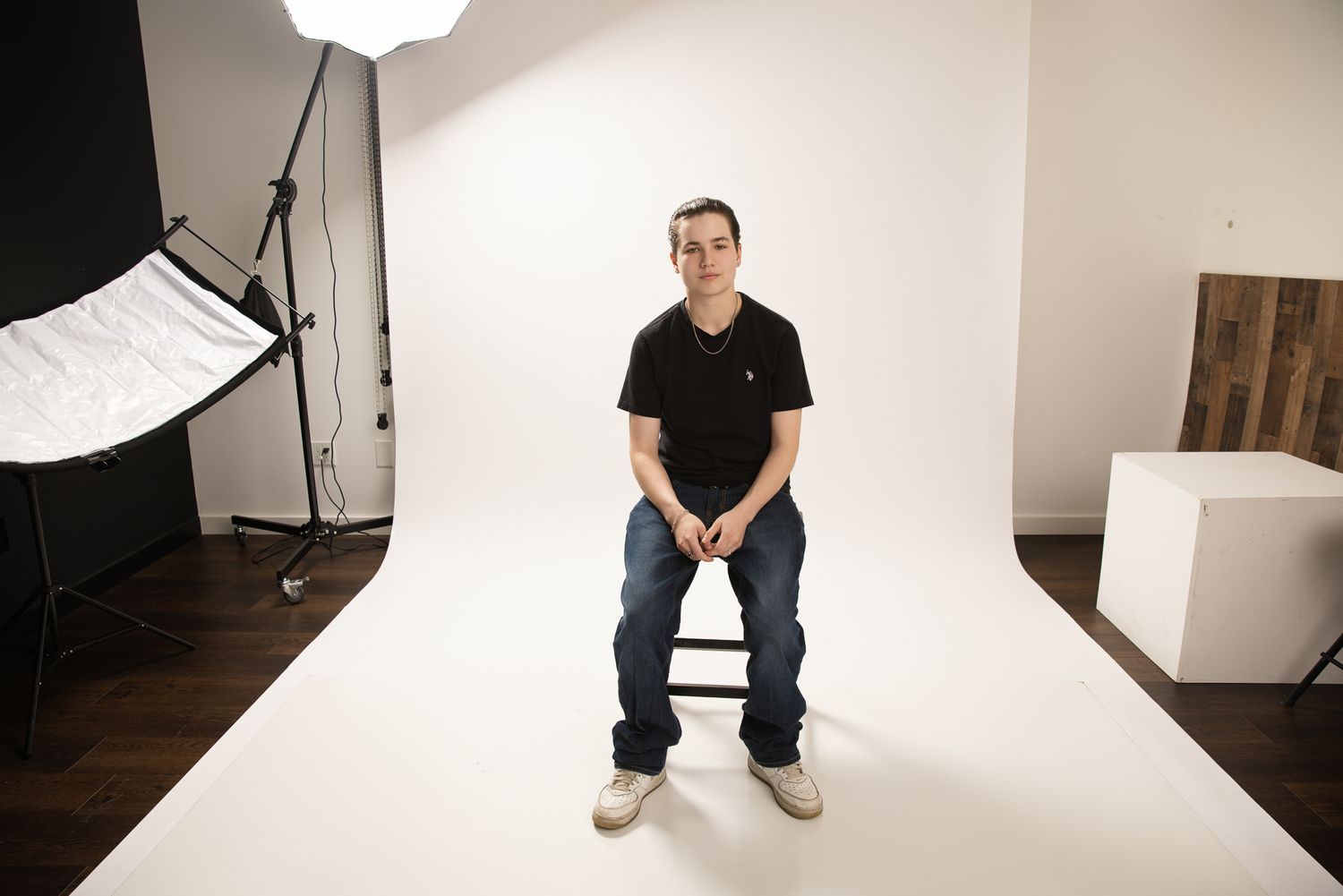 Young man in black polo shirt sitting on stool against white seamless backdrop in professional photography studio.