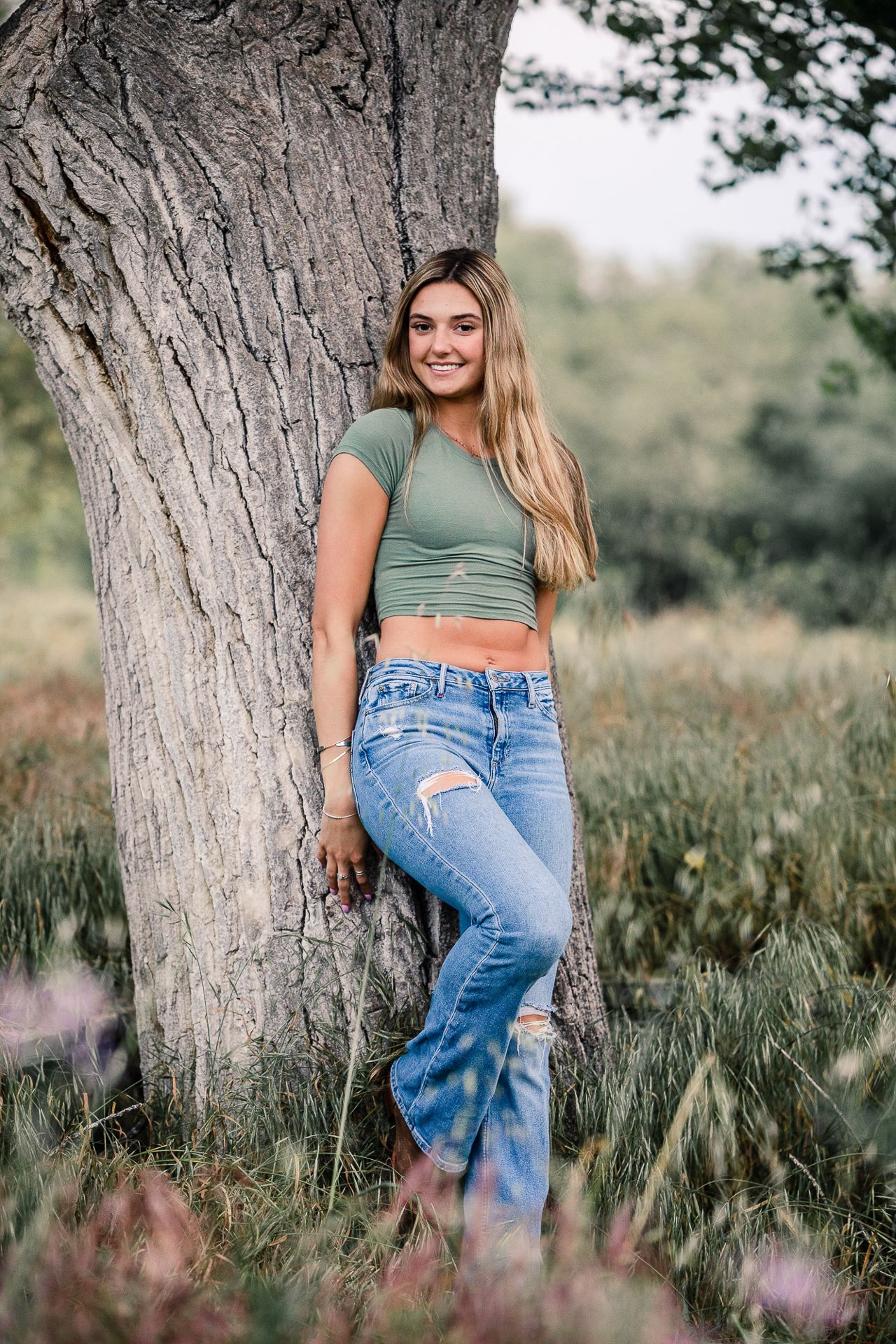 Smiling blonde teen girl in green crop top and ripped flare jeans leaning against large tree outdoors.