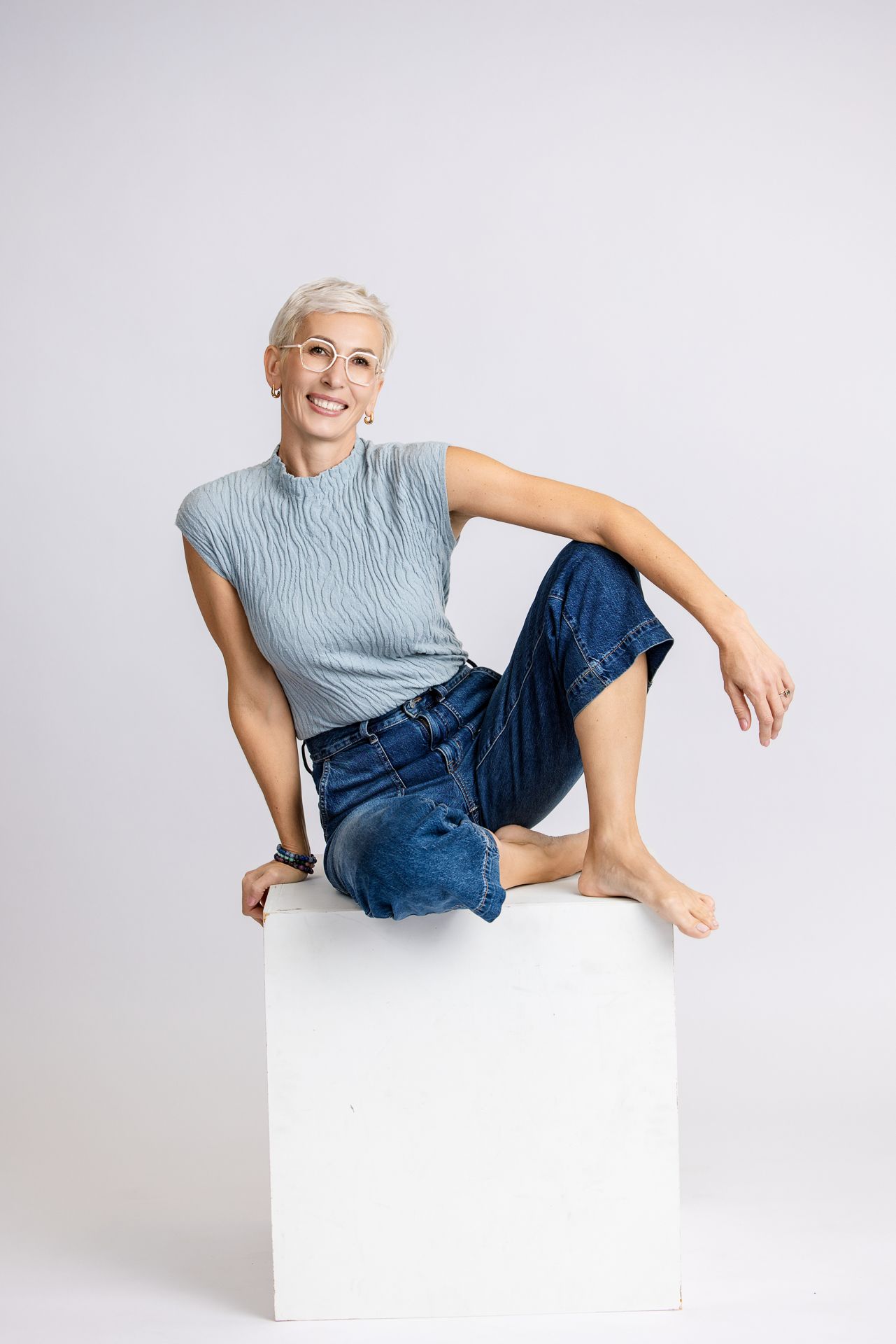 A person with short blonde hair wearing a gray sleeveless top and blue jeans poses elegantly on a white cube.
