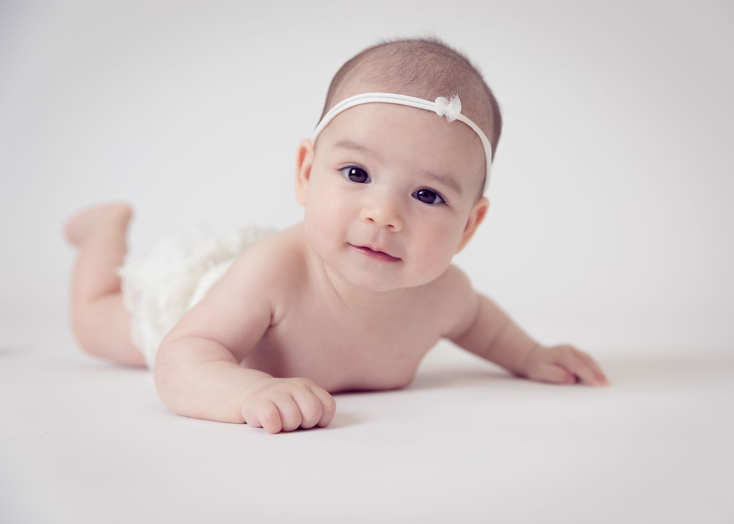 Adorable baby girl in white headband doing tummy time on white background.