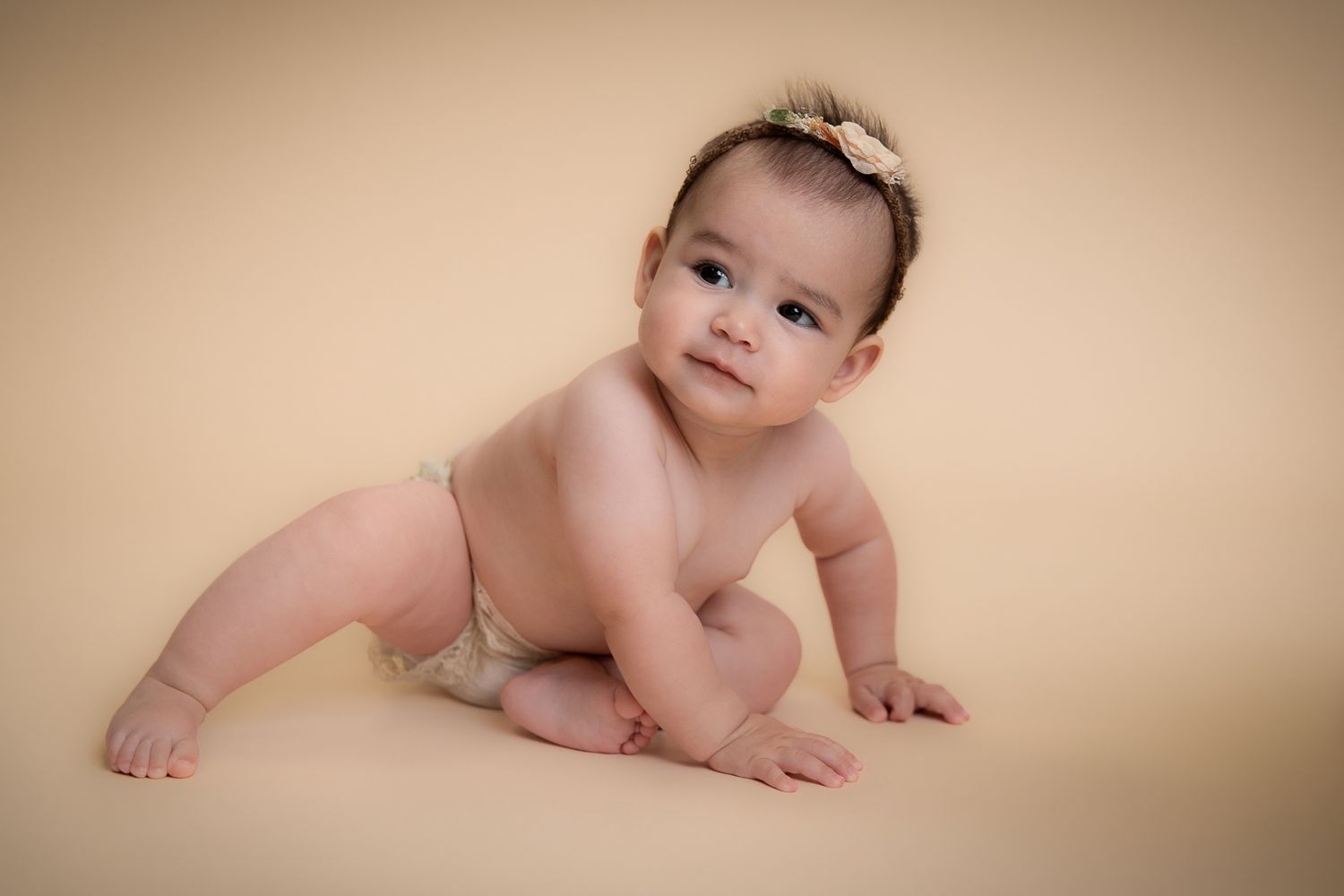 Cute baby in nude diaper crawling on a beige background with a small bow headband.