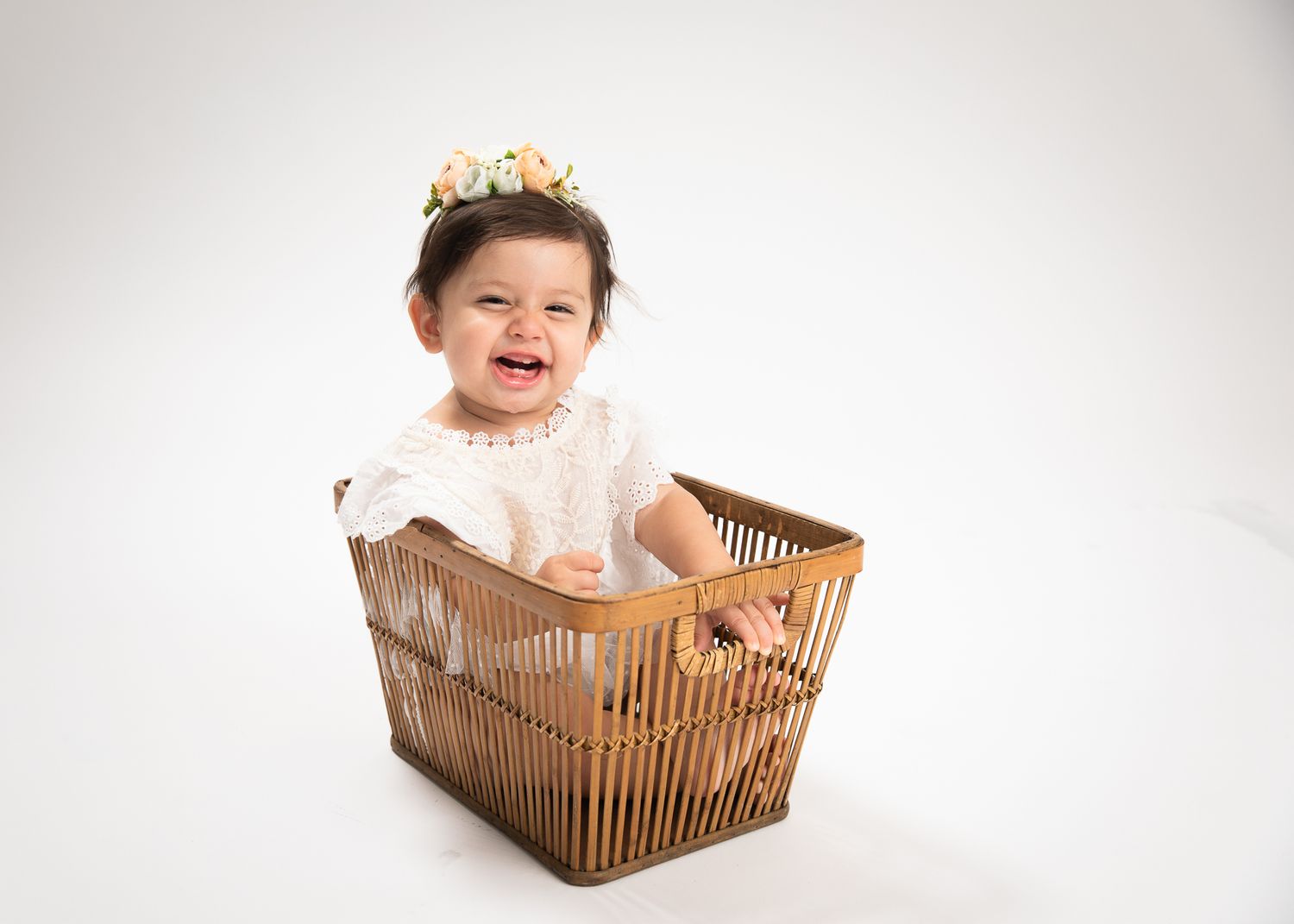 Laughing baby girl in white lace dress and floral crown sitting in a wooden basket.