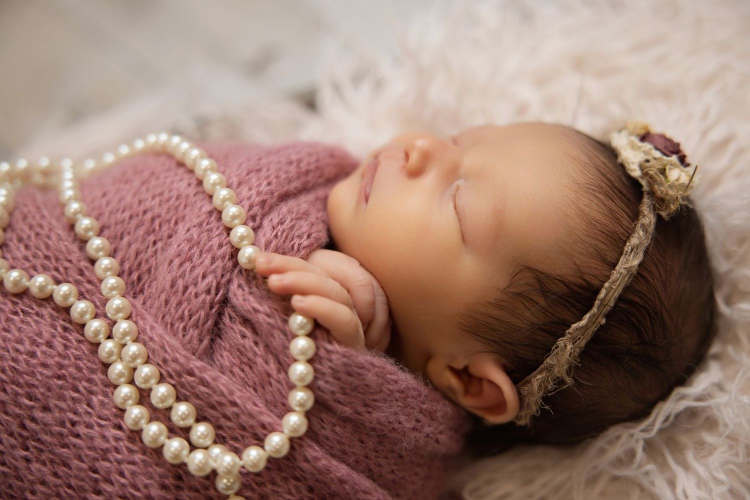 Sleeping newborn baby wrapped in pink knit swaddle wearing pearl necklace and floral crown headband.