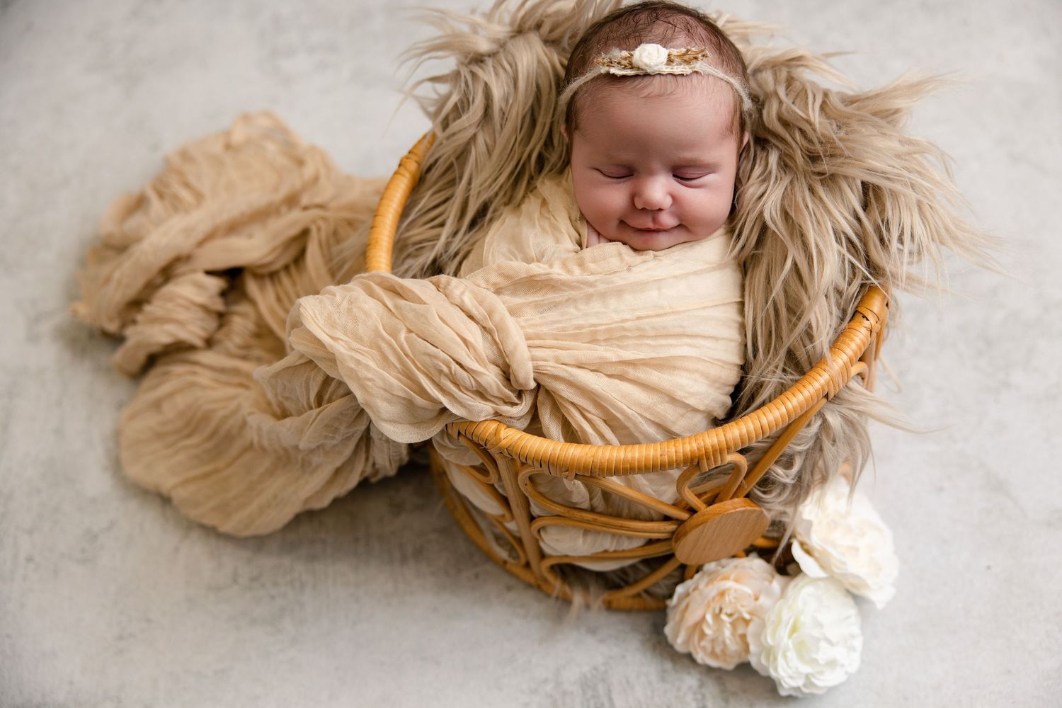Smiling newborn baby wrapped in beige swaddle nestled in wicker basket with fur lining and floral headband.