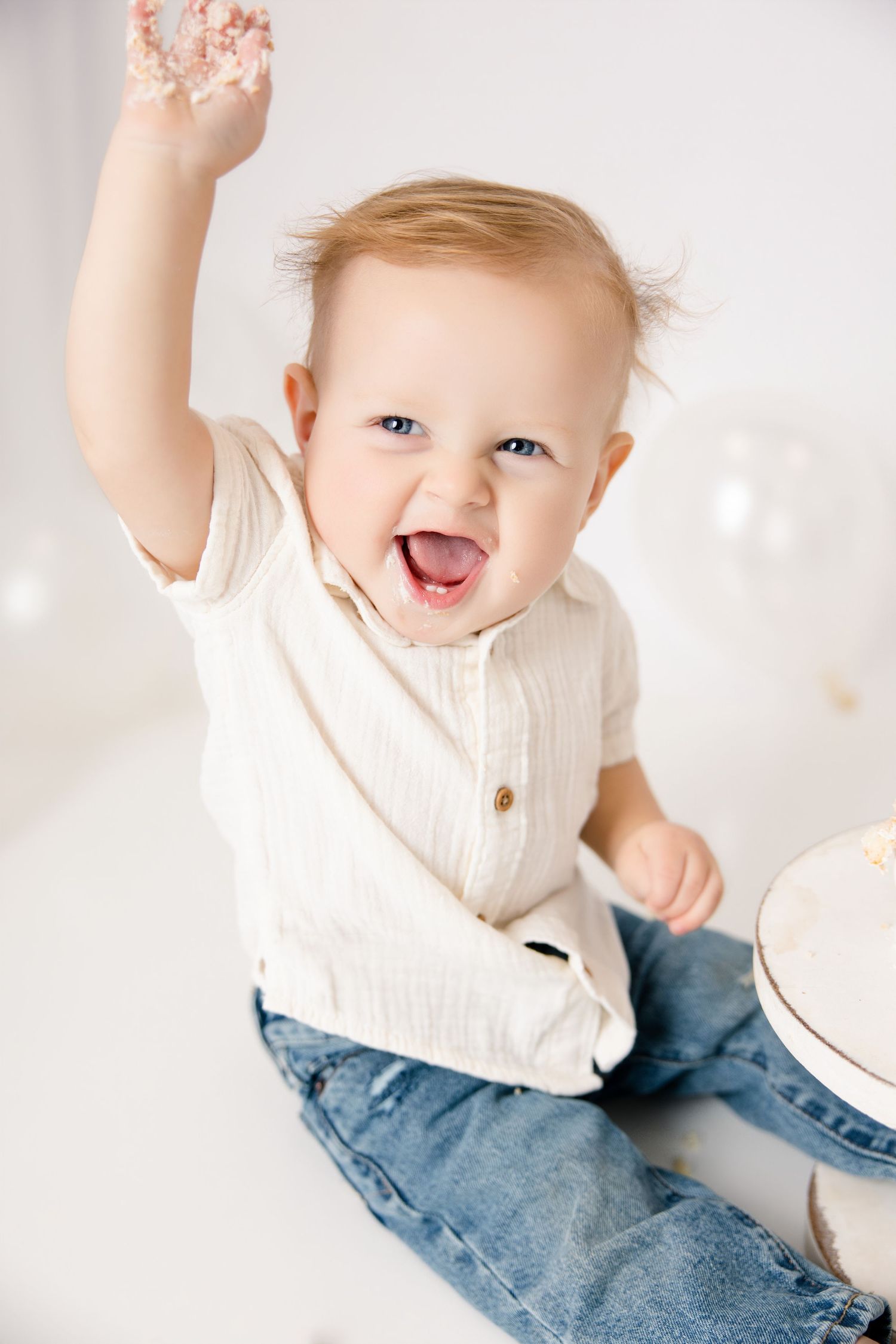 Happy baby celebrating first birthday, smashing white cake with frosting-covered hand, wearing jeans and cream top.