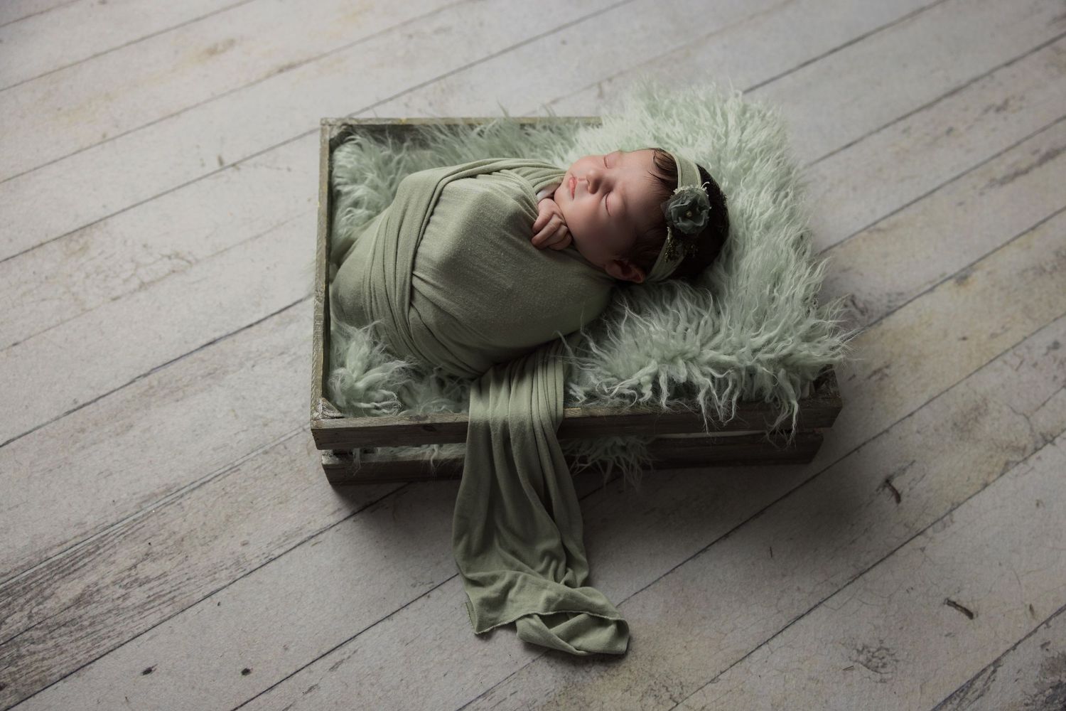 Newborn baby swaddled in sage green wrap resting in a rustic wooden crate with fluffy green fur on white wood floor.