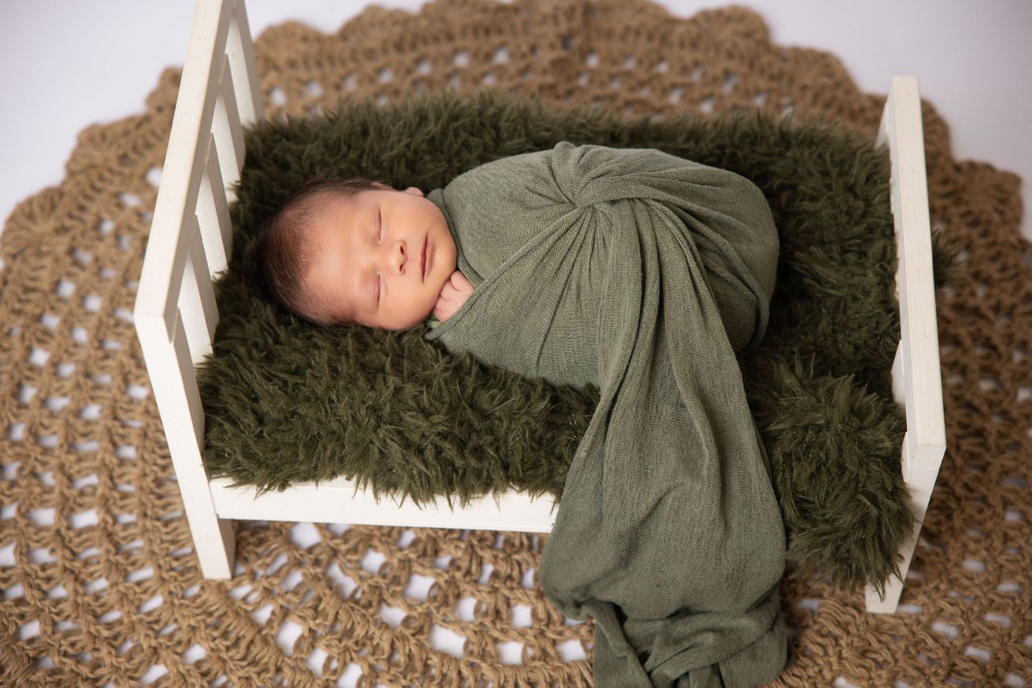 Newborn baby wrapped in green swaddle sleeping on fluffy moss fur on white mini bed prop.
