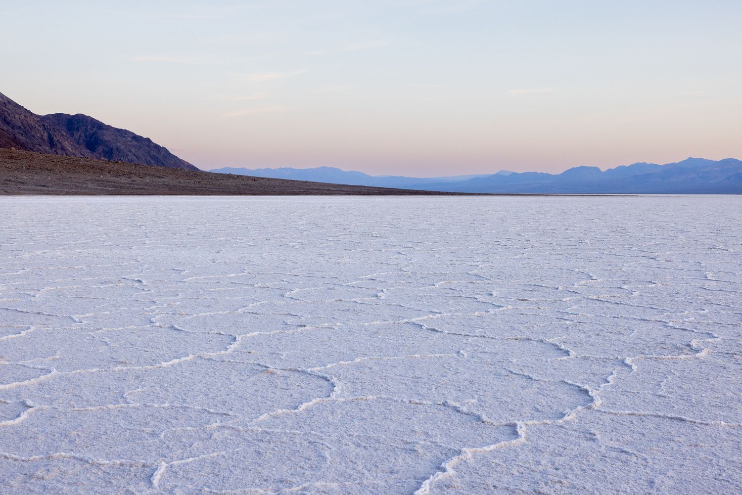 Vast salt flats stretch across Death Valley at dusk, with mountain silhouettes and a pastel sky in the background.