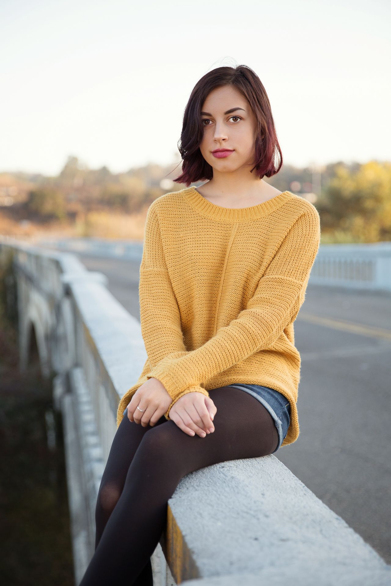 Young woman in yellow knit sweater and shorts sits on bridge railing on autumn day.