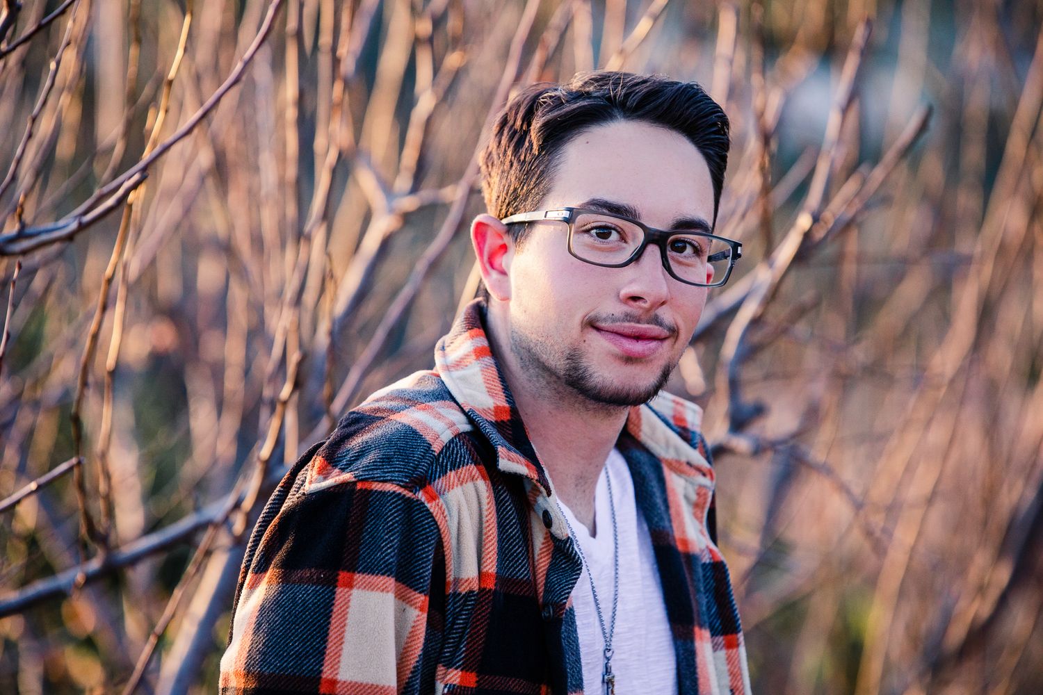 Young man with glasses in plaid shirt posing outdoors among bare branches.