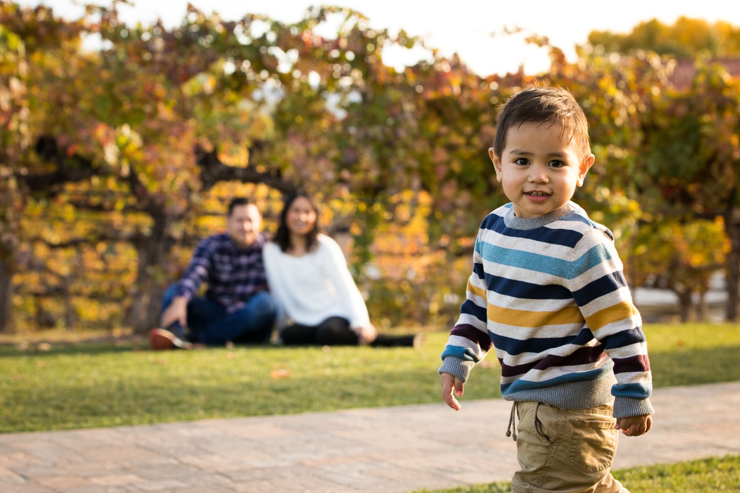 A toddler boy in a striped shirt runs on a path while his parents relax on grass behind him.