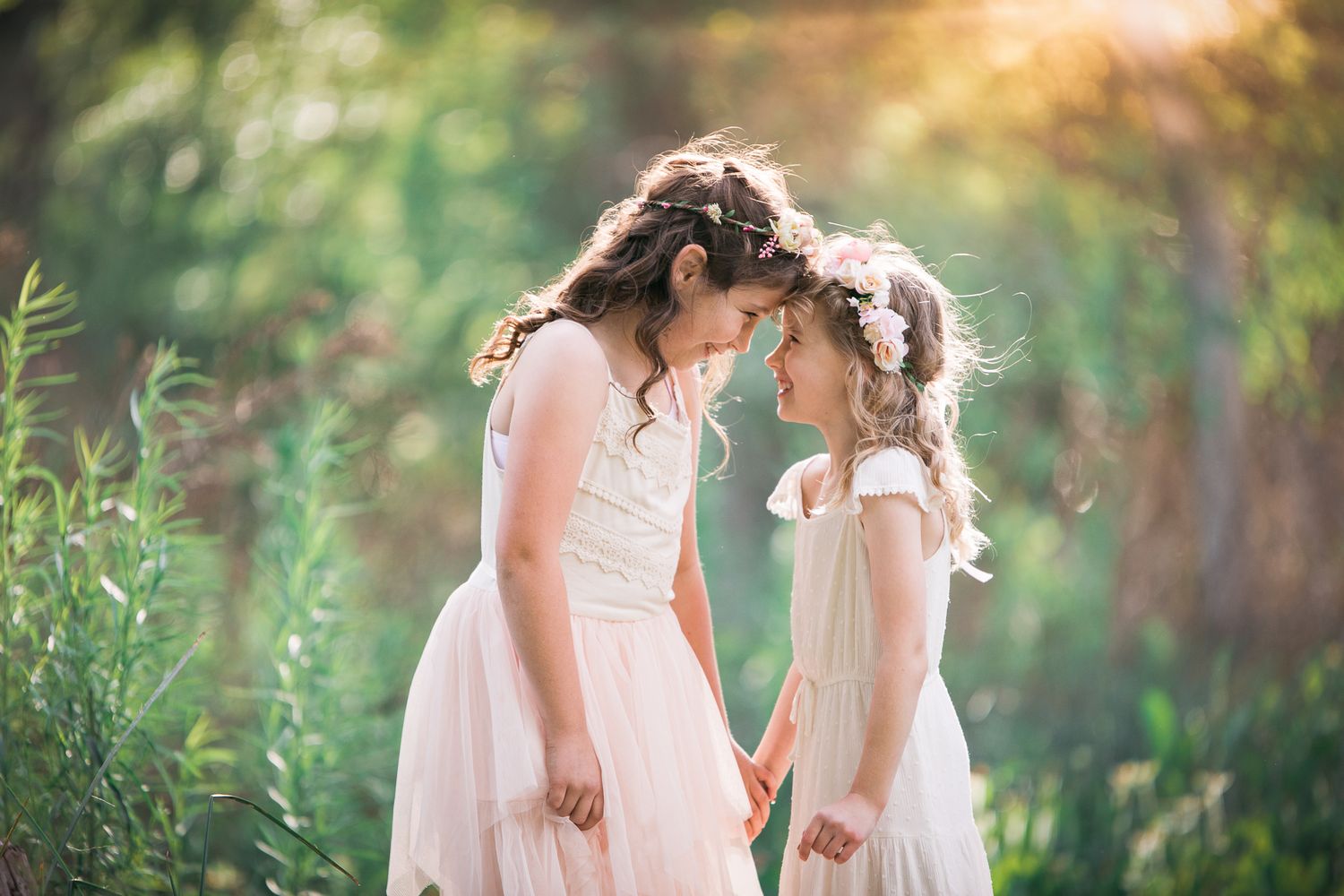Two young girls in white dresses touching foreheads in a sunlit preserve in Fallbrook
