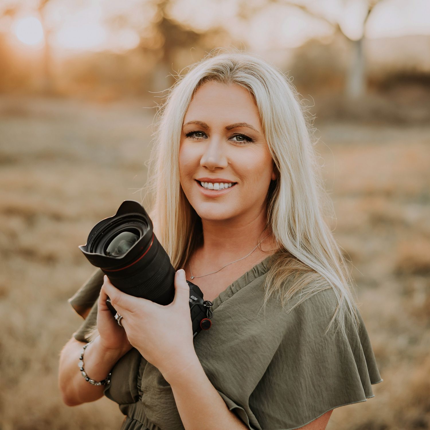 Professional photographer in olive green shirt holding camera outdoors during golden hour sunset.