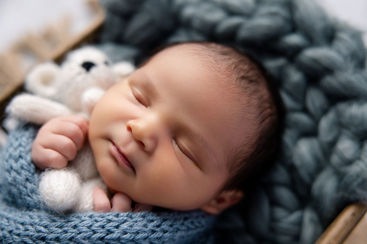Sleeping newborn baby wrapped in blue knit blanket, smiling peacefully while holding a tiny white teddy bear.