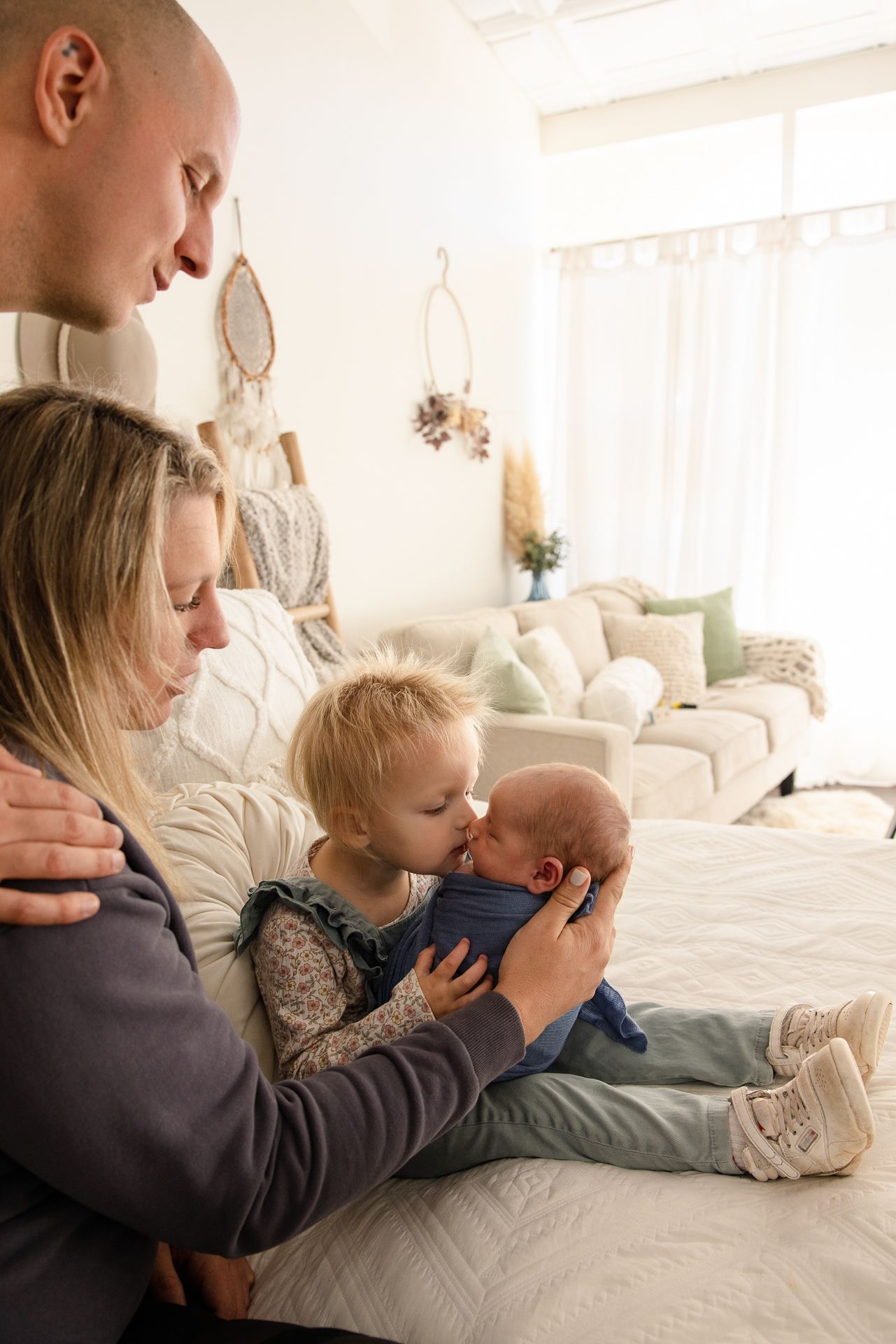 Family with toddler kissing newborn baby on bed in bright boho nursery room.