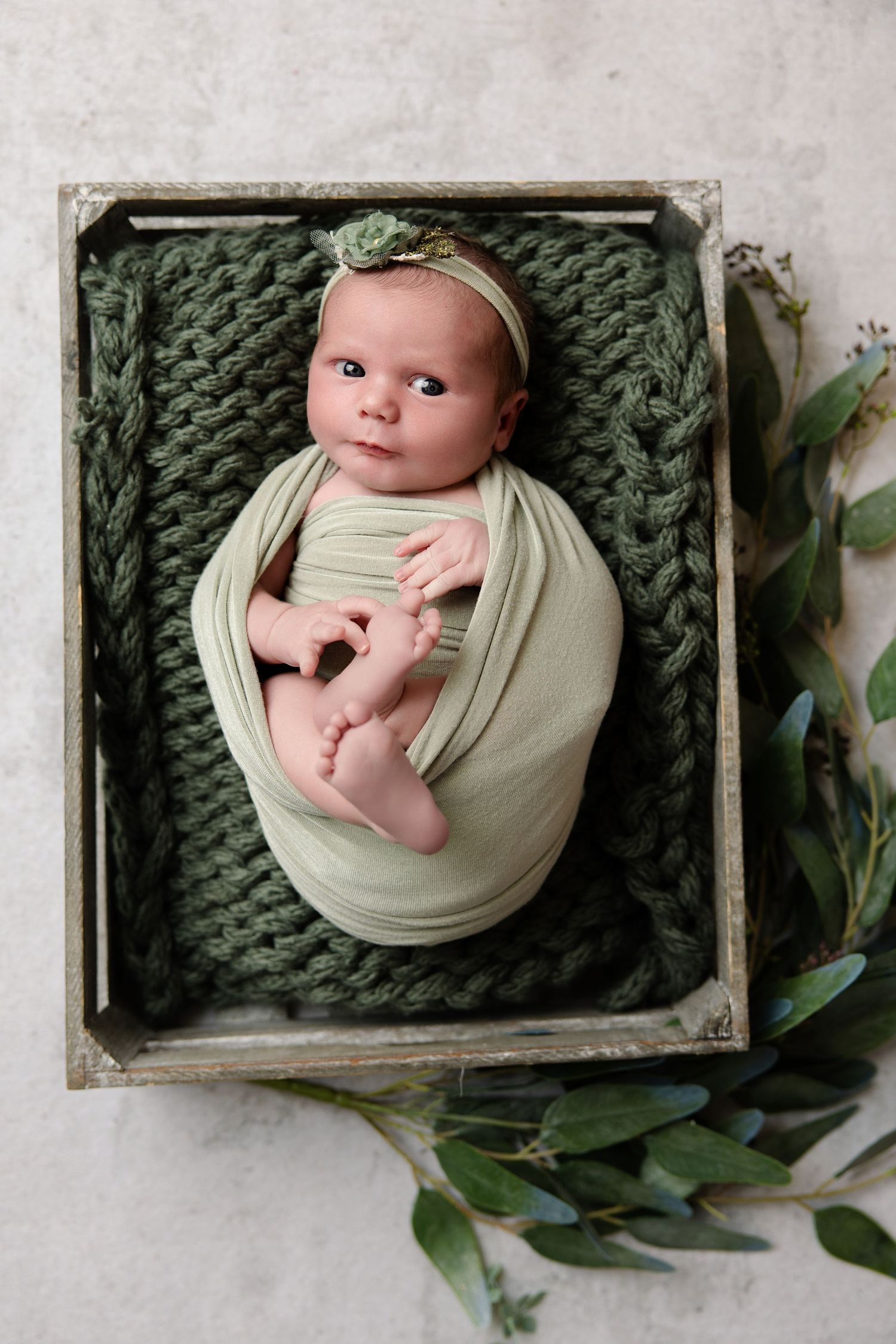 Newborn baby swaddled in sage green wrap, lying in wooden crate with knit blanket and floral headband, surrounded by greenery.