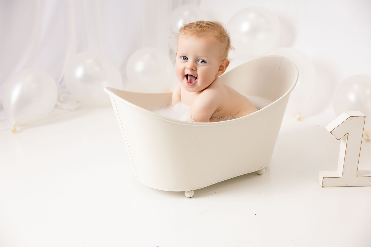 Happy baby sitting in a white mini bathtub surrounded by balloons next to a number one decoration.