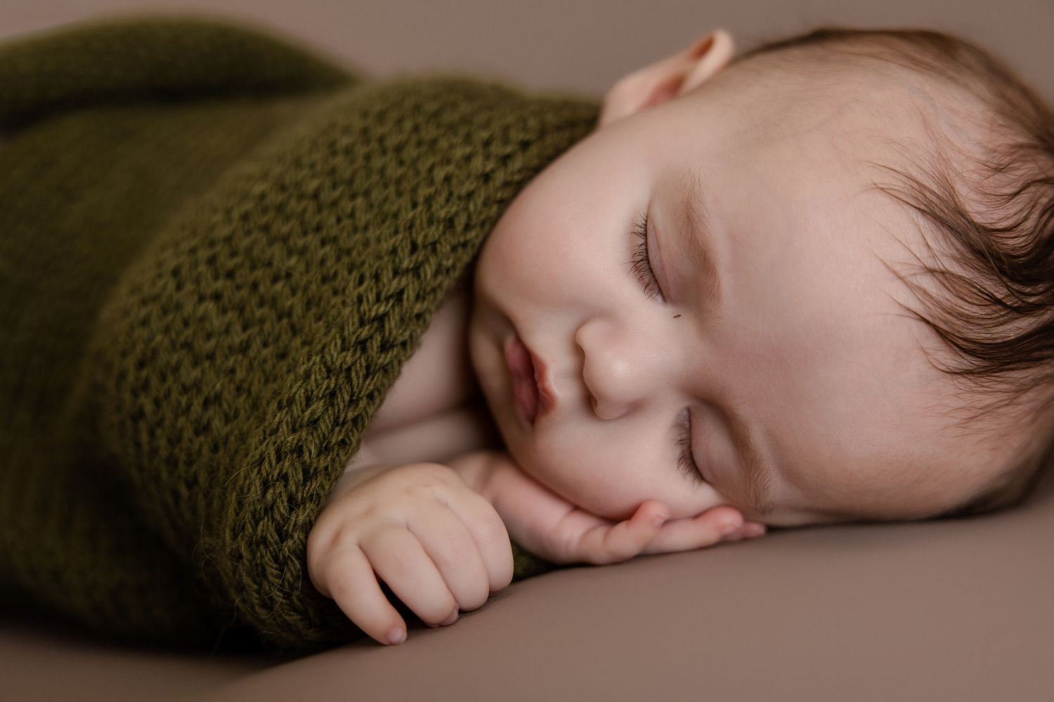 Peaceful newborn baby sleeping wrapped in a soft olive green knit blanket on a neutral taupe background.
