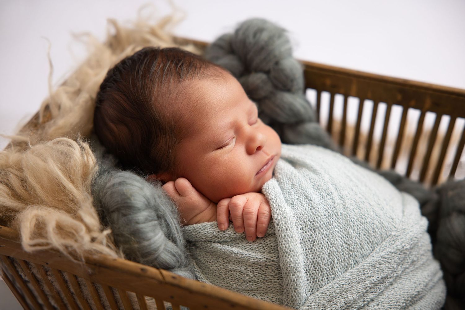 Peaceful newborn baby sleeping in a rustic wooden basket wrapped in a soft gray knit blanket with wool accents.