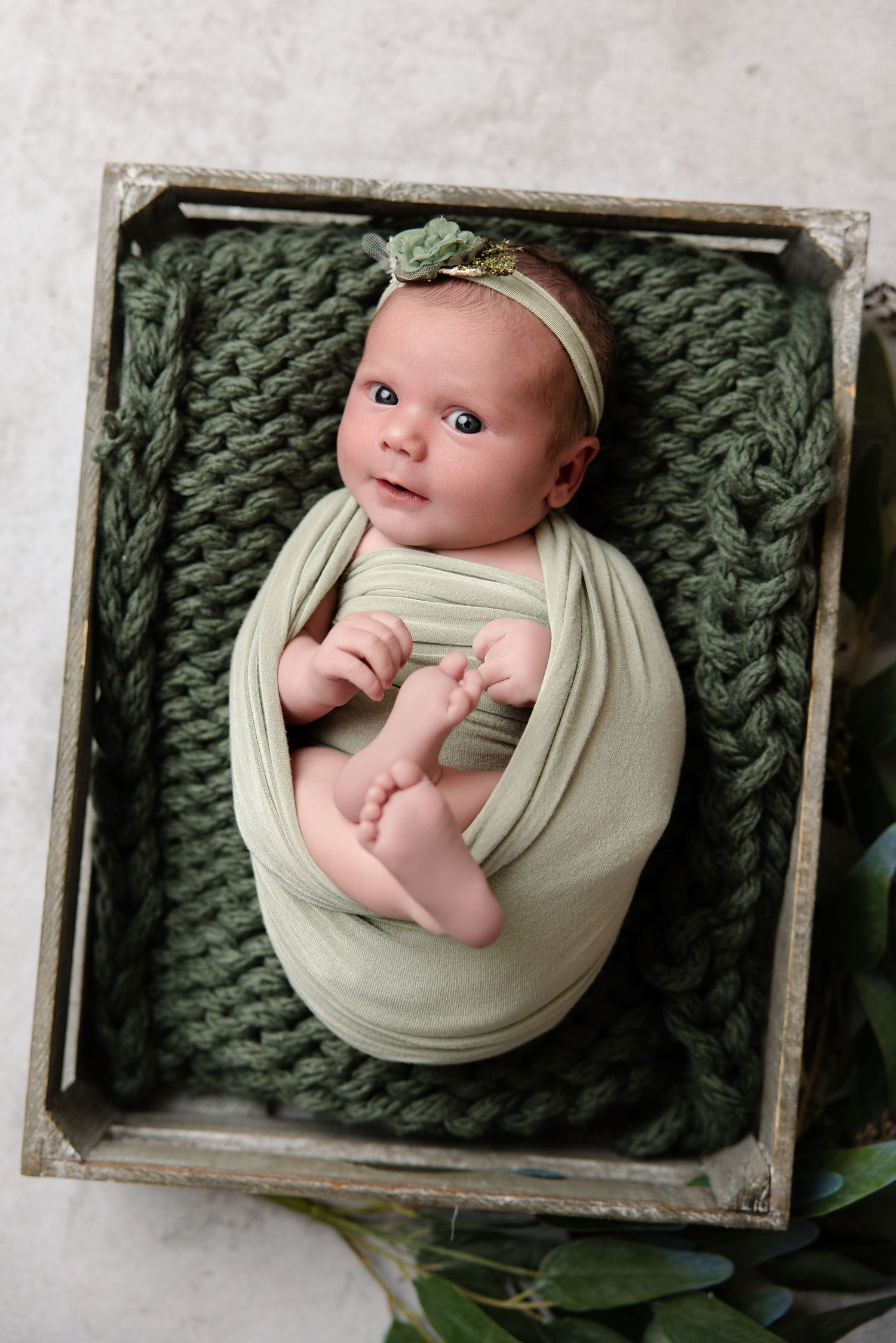 Newborn baby wrapped in light green swaddle with floral headband, resting on dark green knit blanket in wooden crate.