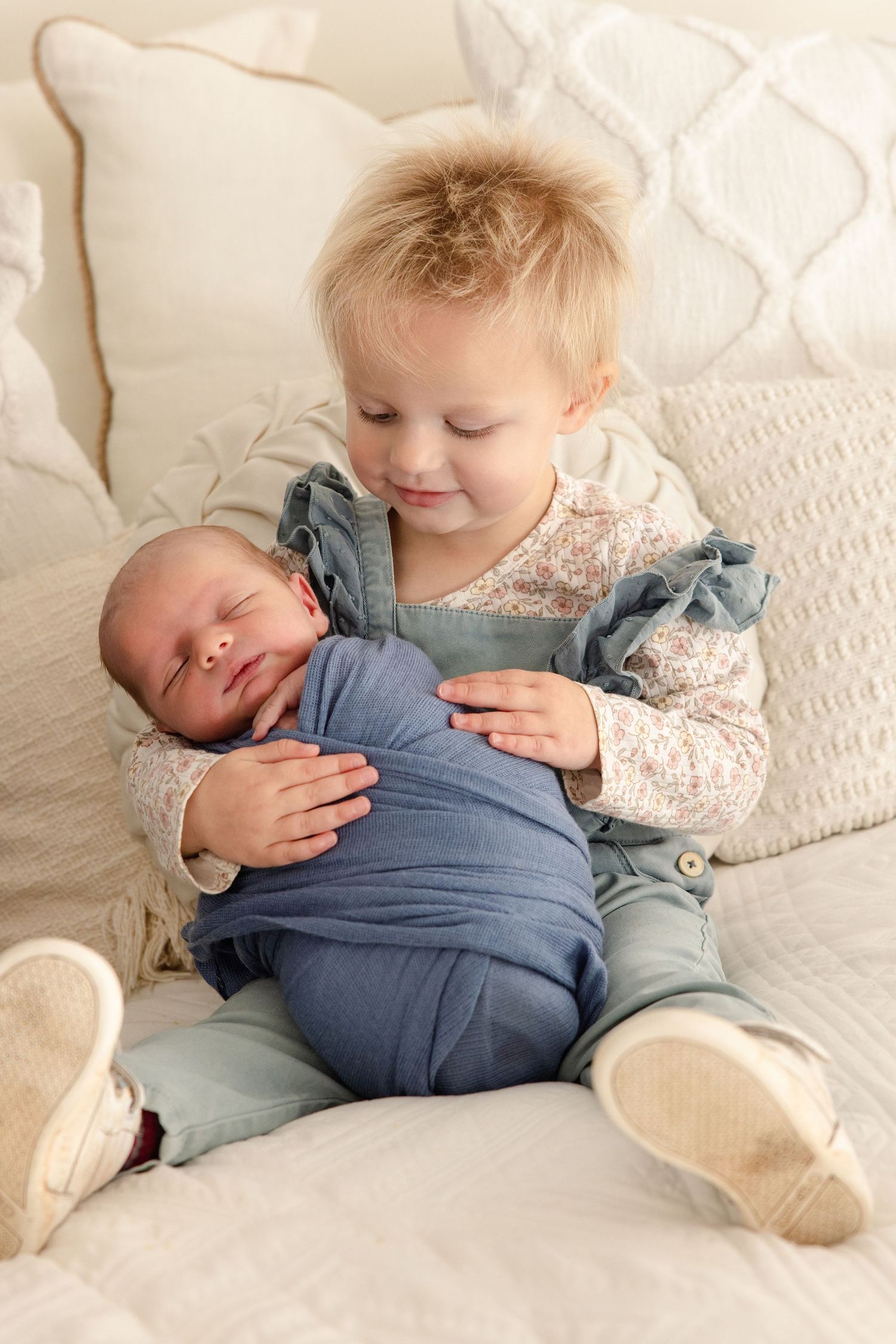 Toddler girl in teal overalls smiles while holding sleeping newborn baby wrapped in blue swaddle on white bed.