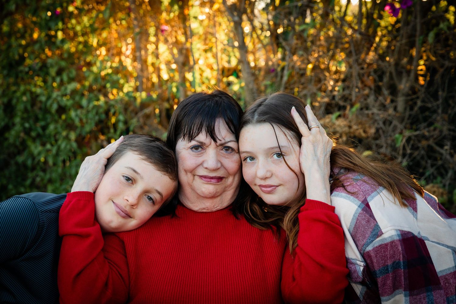A woman in a red sweater smiles warmly with two children leaning on her shoulders outdoors in autumn.