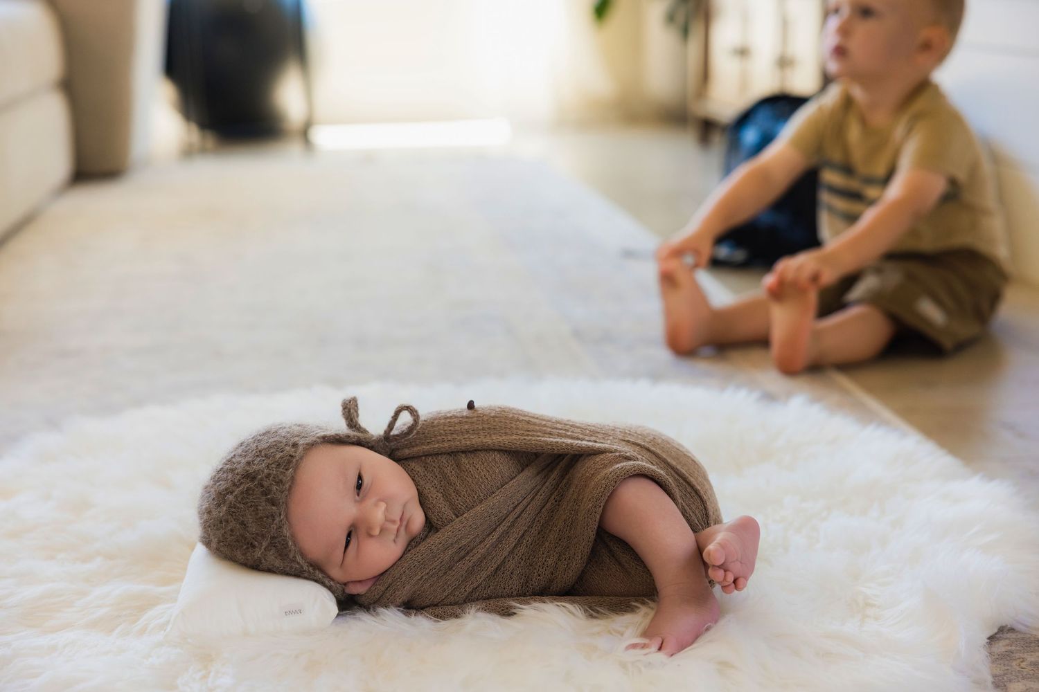 Newborn baby swaddled in brown wrap with antler hat lying on white fur rug beside toddler boy indoors.